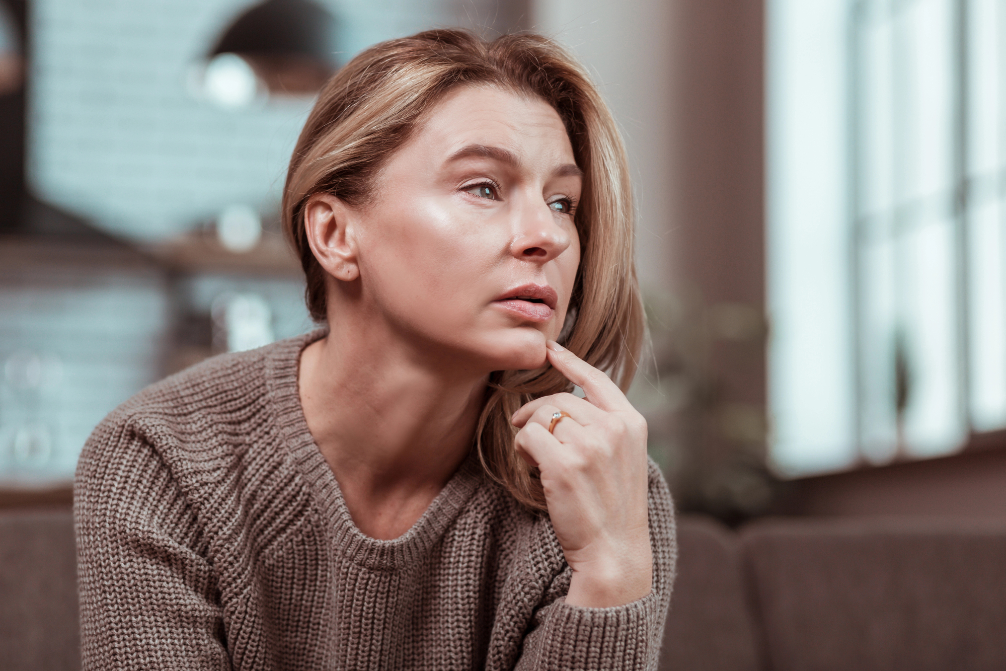 A woman with light hair sits pensively on a couch, resting her chin on her hand. She is wearing a light brown sweater and looking slightly to the side. The background is softly blurred, showing a cozy indoor setting with natural light coming through the windows.