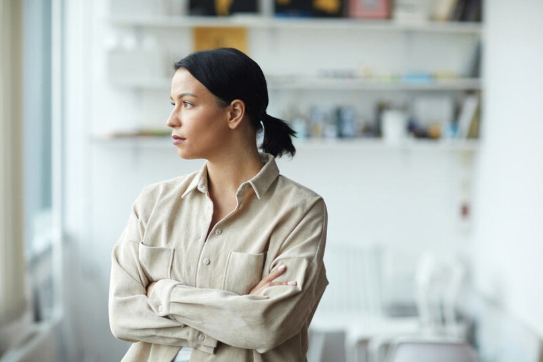 A woman with her hair tied back is standing indoors with her arms crossed, looking thoughtfully out of the window. She is wearing a beige button-up shirt. There are shelves with various items and books blurred in the background.