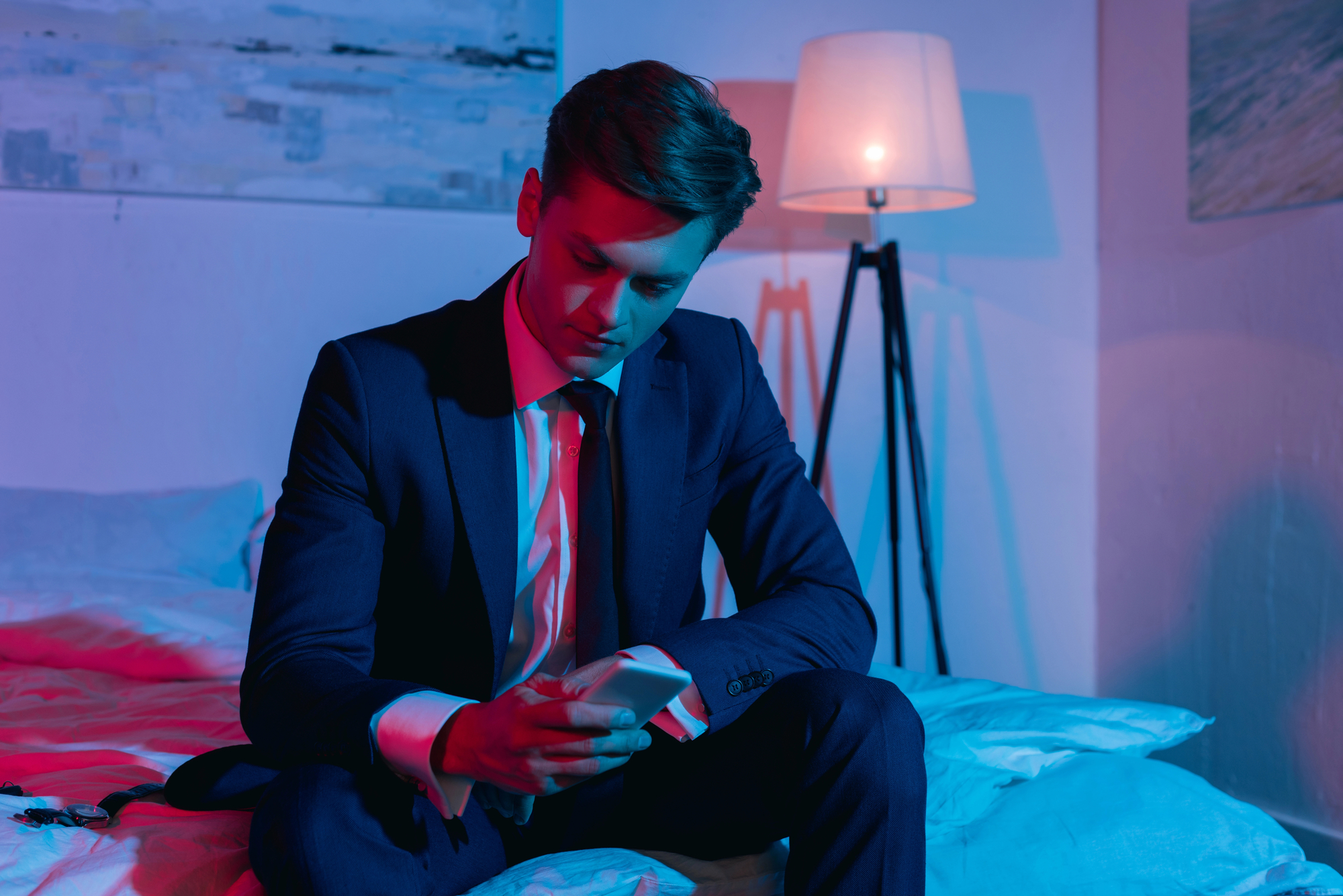 A man in a suit sits on a bed in a dimly lit room, illuminated by a combination of blue and pink lights. He is looking down at his smartphone, giving an impression of deep focus or contemplation. A floor lamp glows softly in the background.