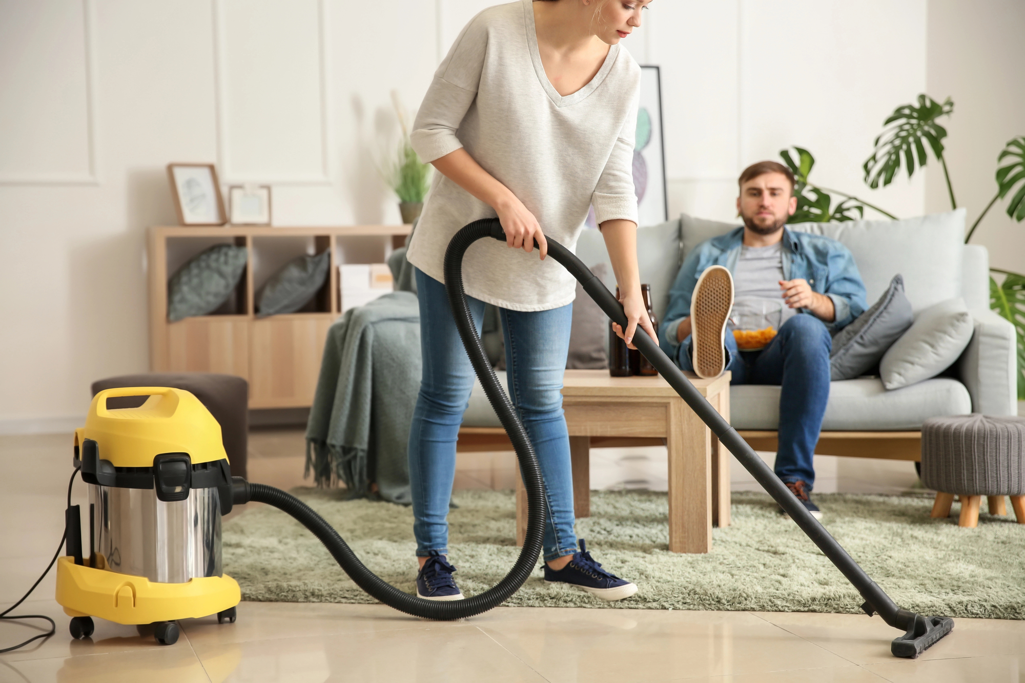 A woman in a casual outfit is vacuuming the floor with a yellow and silver vacuum cleaner in a living room. In the background, a man in a denim jacket and jeans is sitting on a light blue sofa, eating snacks and watching her. The room is modern and bright.
