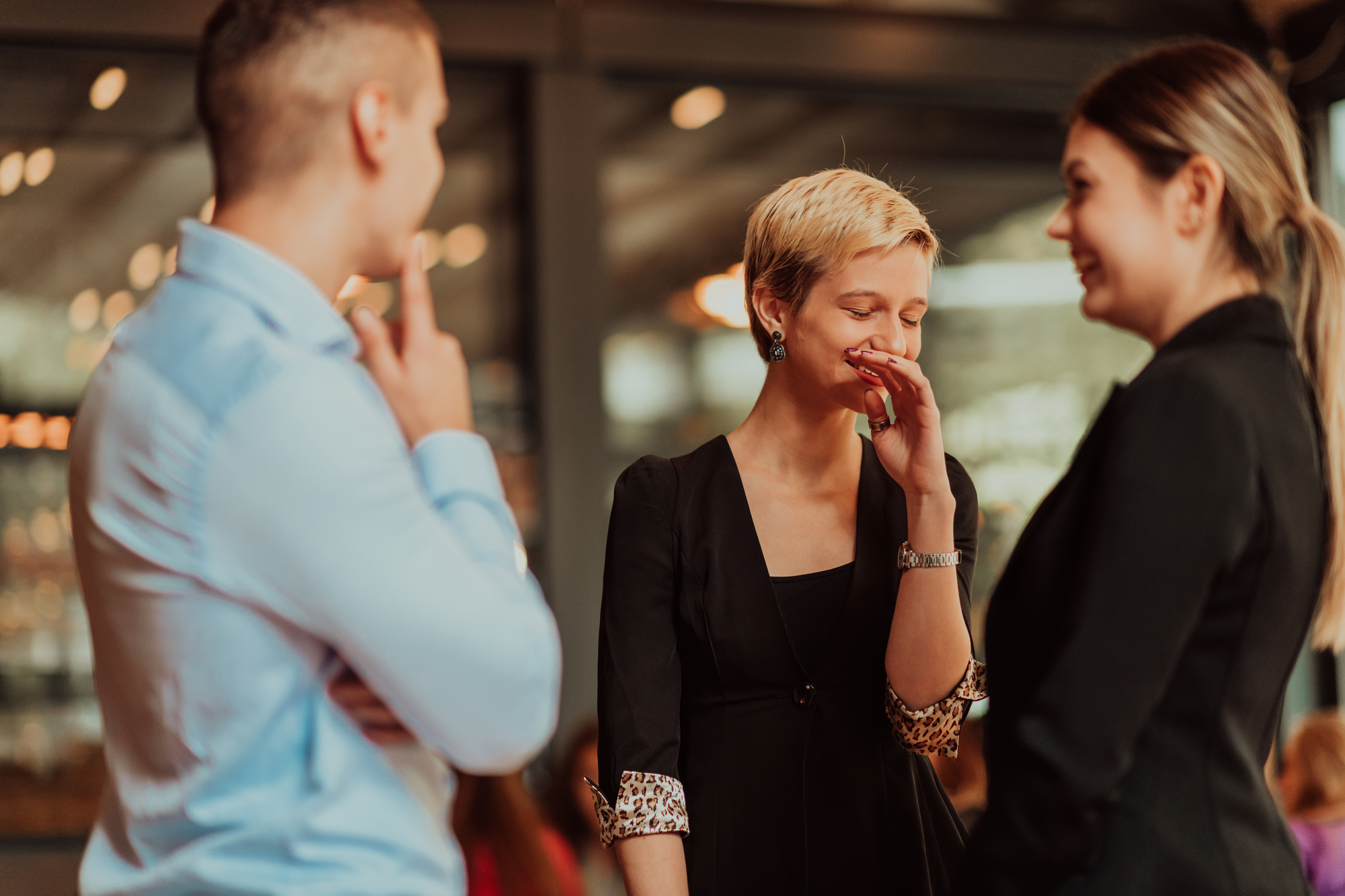 Three people are standing and engaging in a lively conversation. A person in a black outfit is laughing with a hand covering their mouth, while another person in a black outfit is smiling. A third person in a blue shirt is observing and touching their chin.