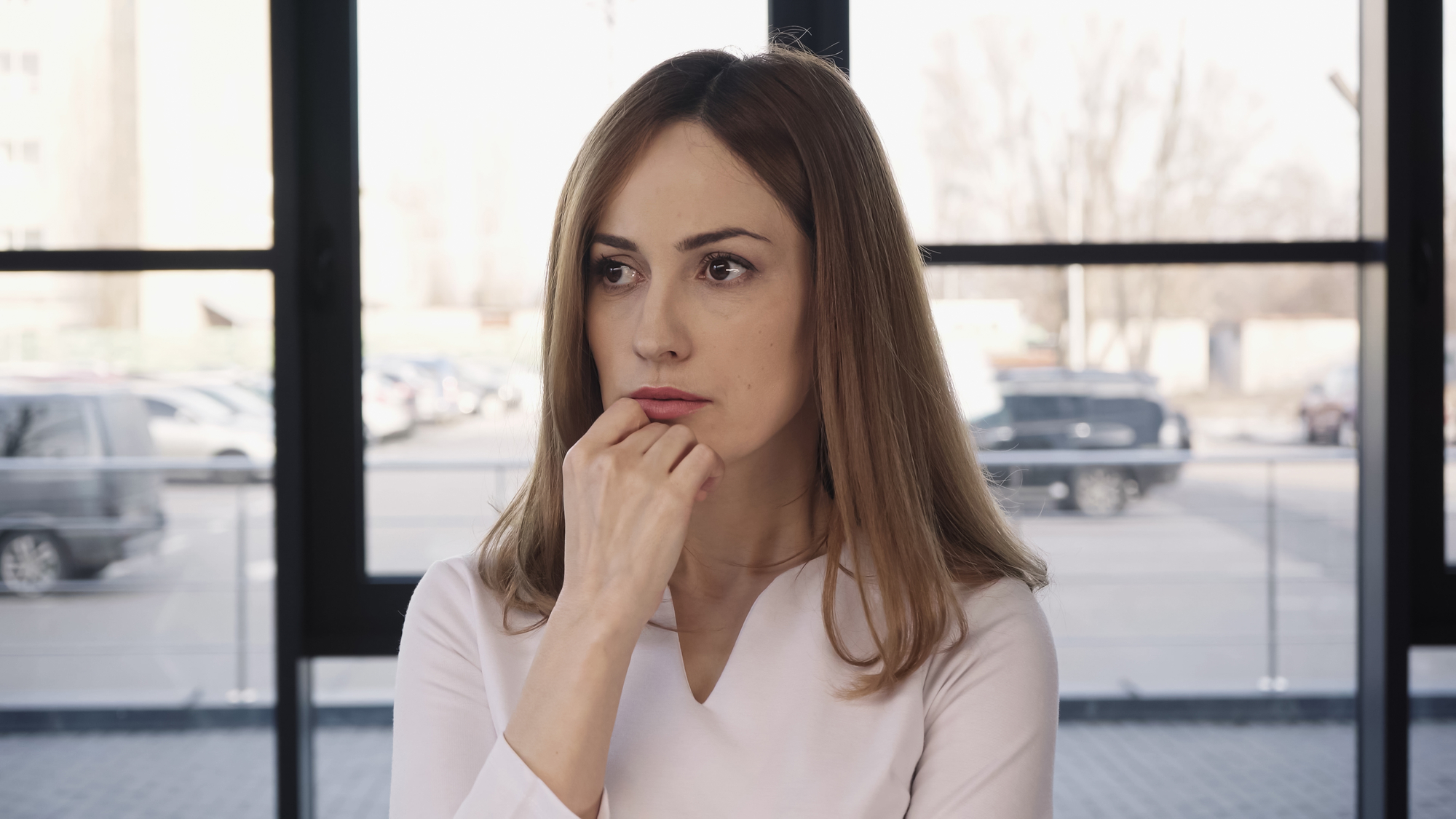 A woman with long brown hair stands indoors in front of large windows. She is wearing a white top and has a thoughtful expression, with one hand resting near her mouth. Outside the windows, a parking lot and bare trees are visible.