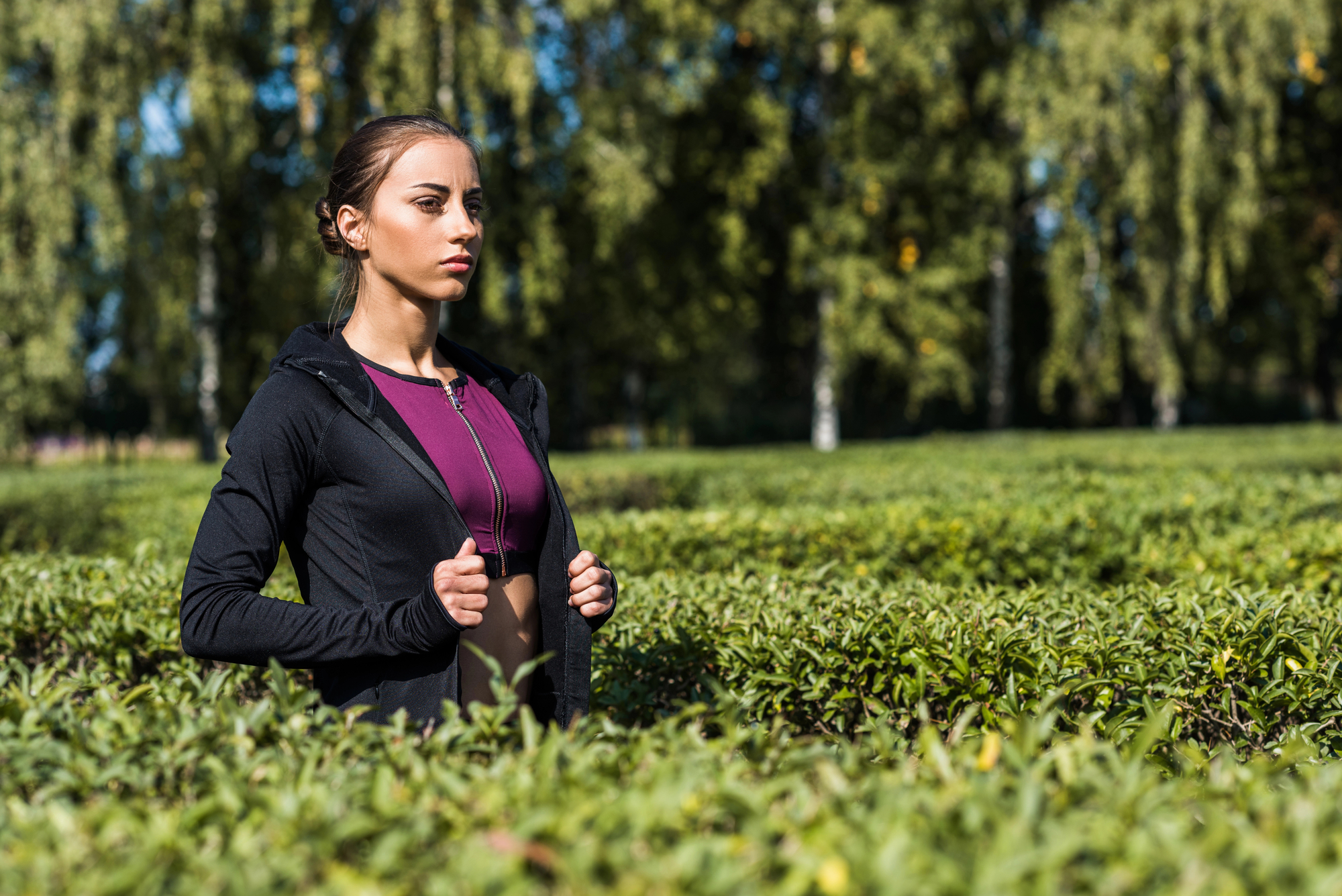 A woman stands in a field of shrubs, zipping up her black hoodie over a purple top. She looks into the distance with a serious expression. The background features green trees with bright sunlight filtering through.