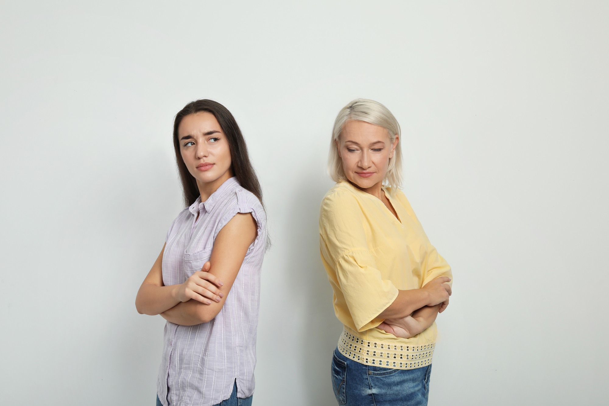 Two women, one with long dark hair and the other with short white hair, stand back-to-back with their arms crossed, both looking slightly annoyed or displeased. They are dressed casually in light-colored tops and blue jeans, standing against a plain white background.