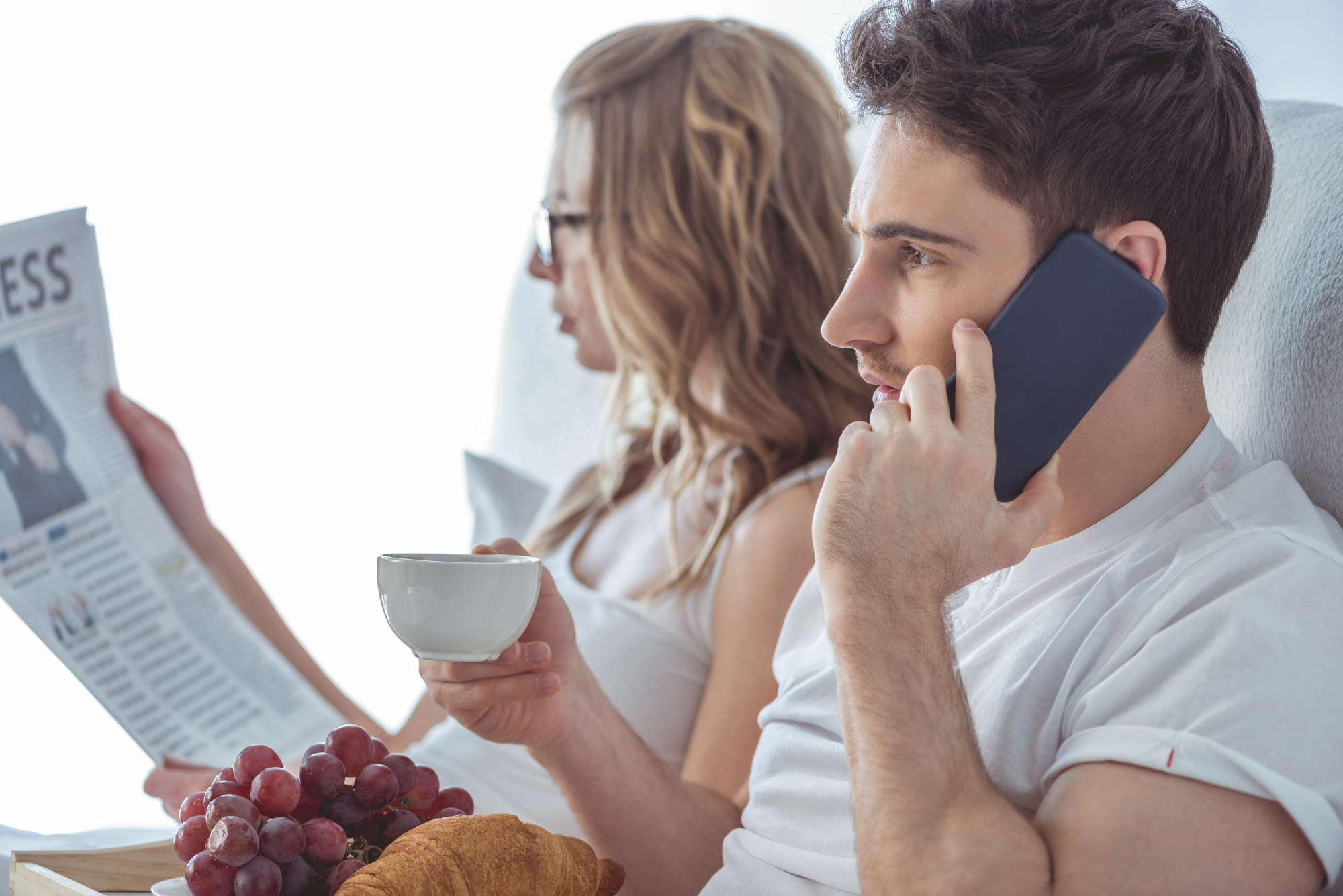 A man and woman are sitting together on a couch. The man is holding a cup, talking on the phone, and a tray with grapes and a croissant is on his lap. The woman, in the background, is reading a newspaper and wearing glasses. Both are casually dressed in white.