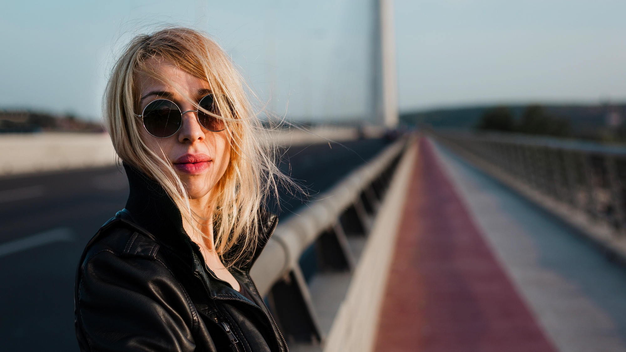 A woman with blonde hair wearing round sunglasses and a black leather jacket stands on a bridge with a blurred background of road and railings. Her hair is slightly windswept, and she gazes into the camera with a neutral expression.