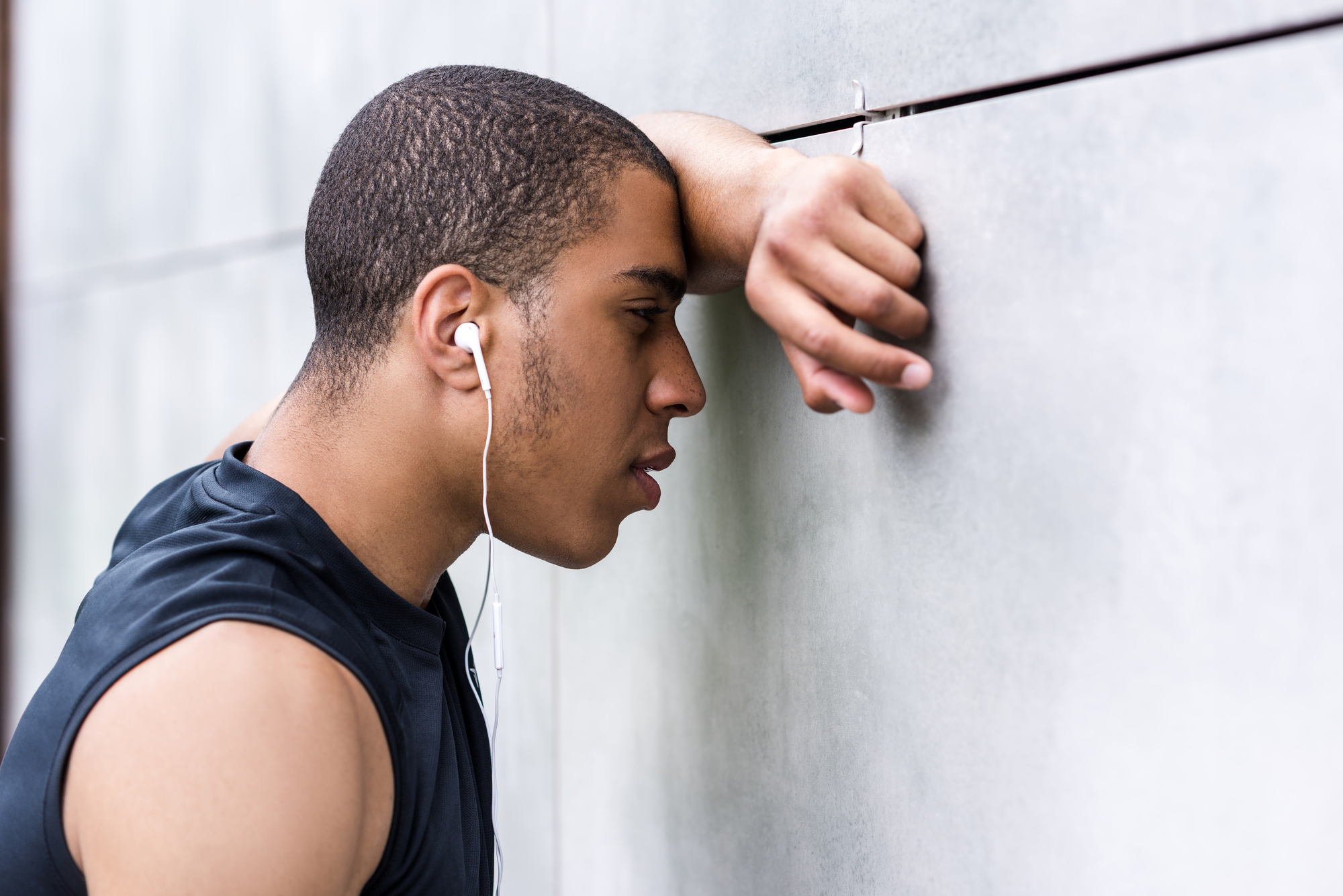 A person wearing a sleeveless black shirt and white earphones leans against a wall, appearing tired or contemplative. Their forehead touches their arm, which is bent at the elbow and rests against the wall.