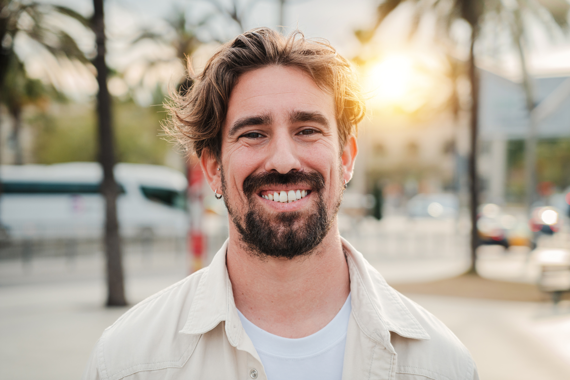 A man with medium-length hair and a beard is smiling outdoors. He is wearing a light-colored jacket and a white shirt. The background features palm trees and the sun setting, creating a warm, pleasant atmosphere.