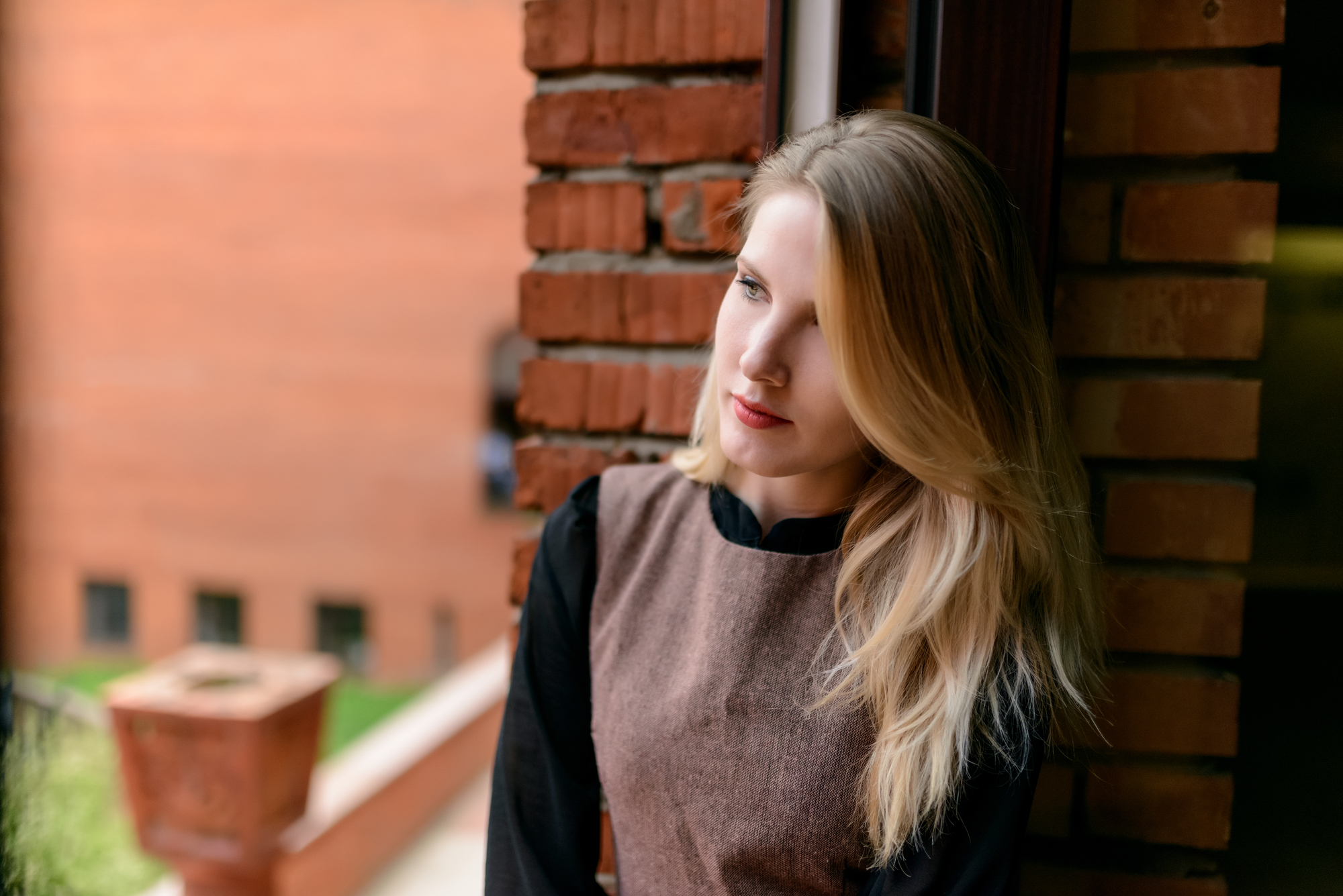 A woman with long, blonde hair and dressed in a brown dress over a black top stands pensively against a brick wall. She gazes off to the side with a thoughtful expression. The background features another brick building and greenery.