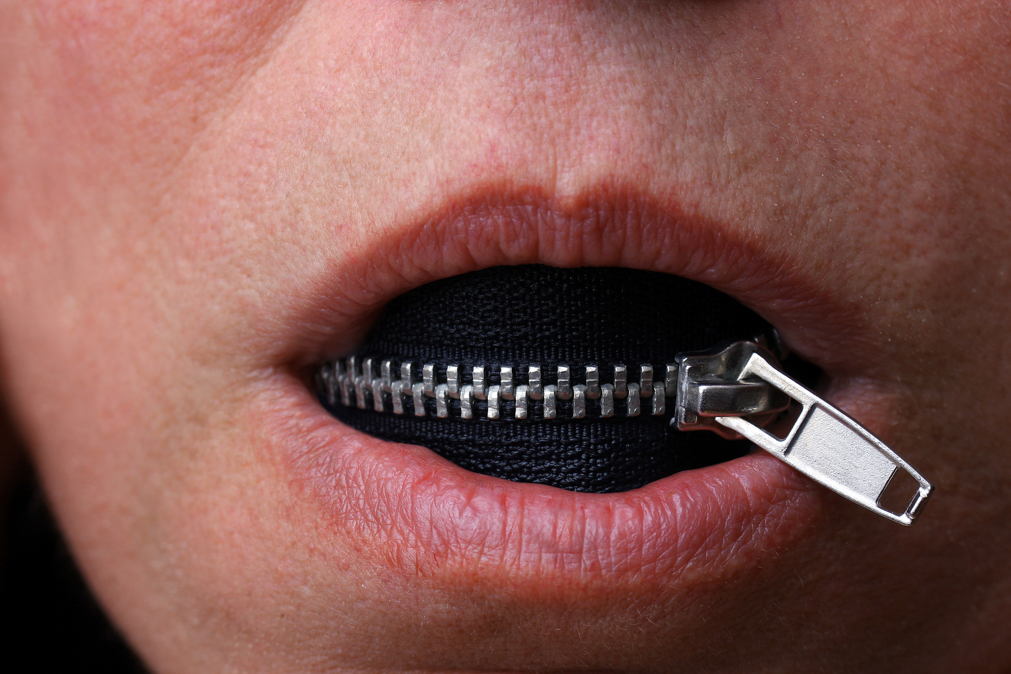 Close-up of a person's lips partially unzipped with a zipper running horizontally across their mouth, revealing black fabric behind the zipper. The image suggests the concept of silence or keeping quiet.