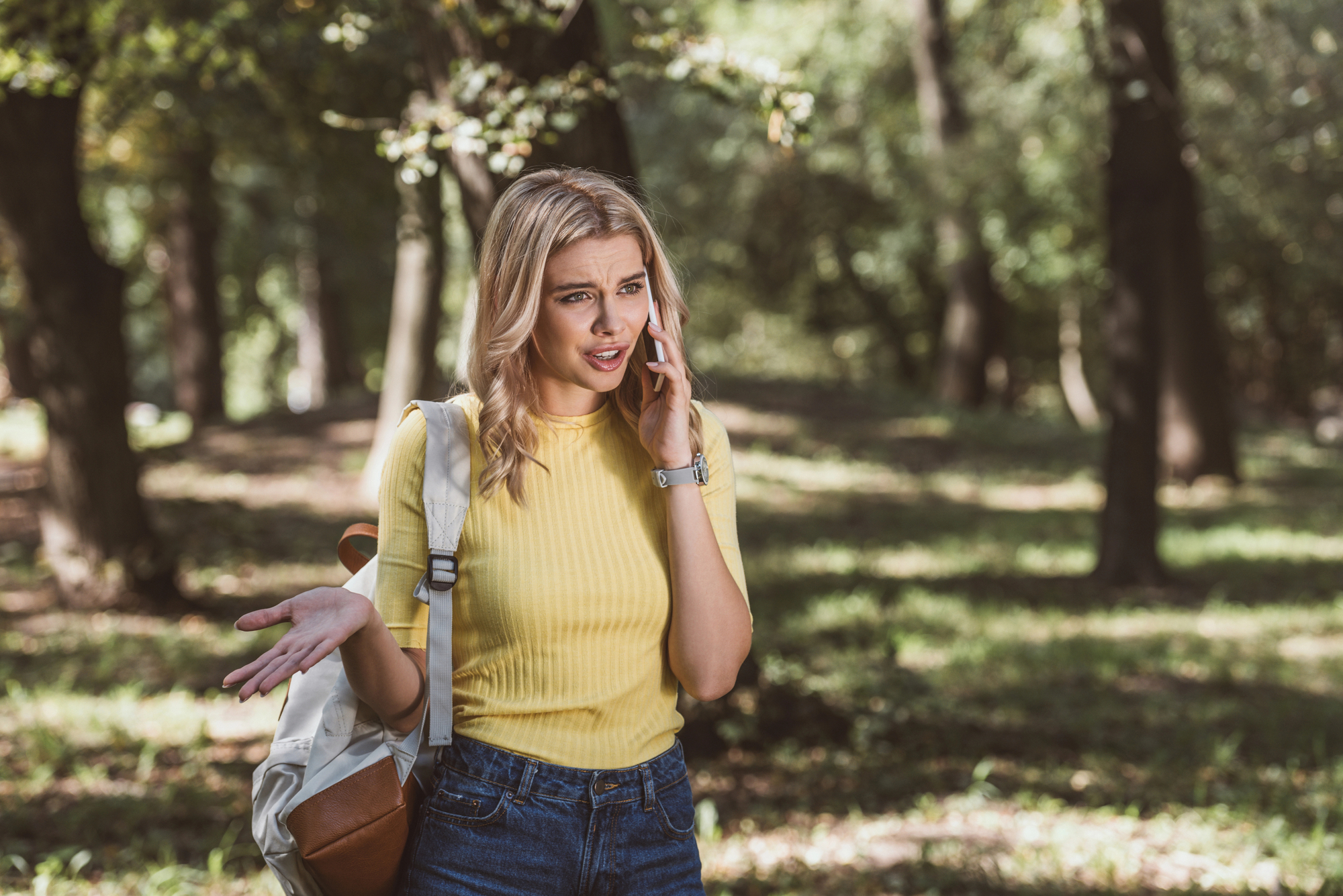 A woman with blonde hair, wearing a yellow shirt and carrying a backpack, stands in a forested area talking on her phone. She looks confused or frustrated, with one hand raised in an expressive gesture. The lush green background suggests it's a sunny day.