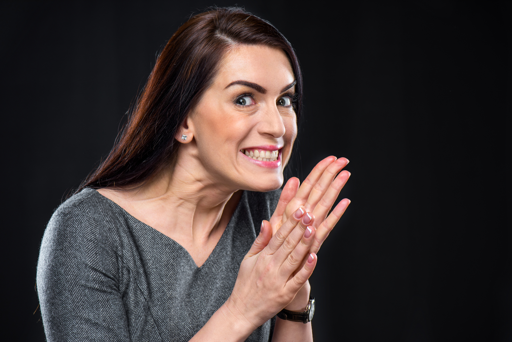 A woman with long dark hair and a gray top smirks while clasping her hands together near her face. She is looking slightly to the side against a dark background, with an expression of anticipation or excitement.