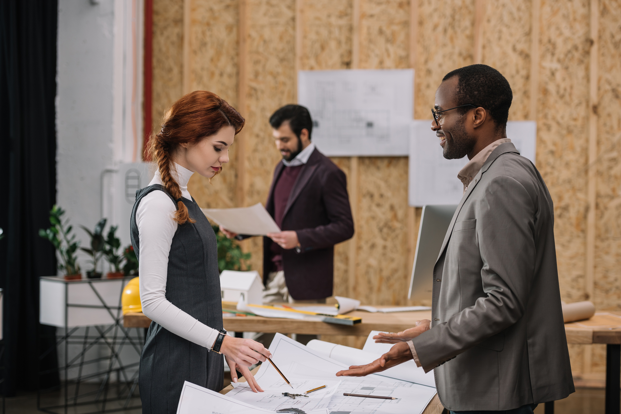 A woman and a man discuss blueprints on a table in an architect's office, with another man reviewing plans in the background. The office has wooden walls and various papers and plans spread out. The trio appears engaged in a collaborative discussion.