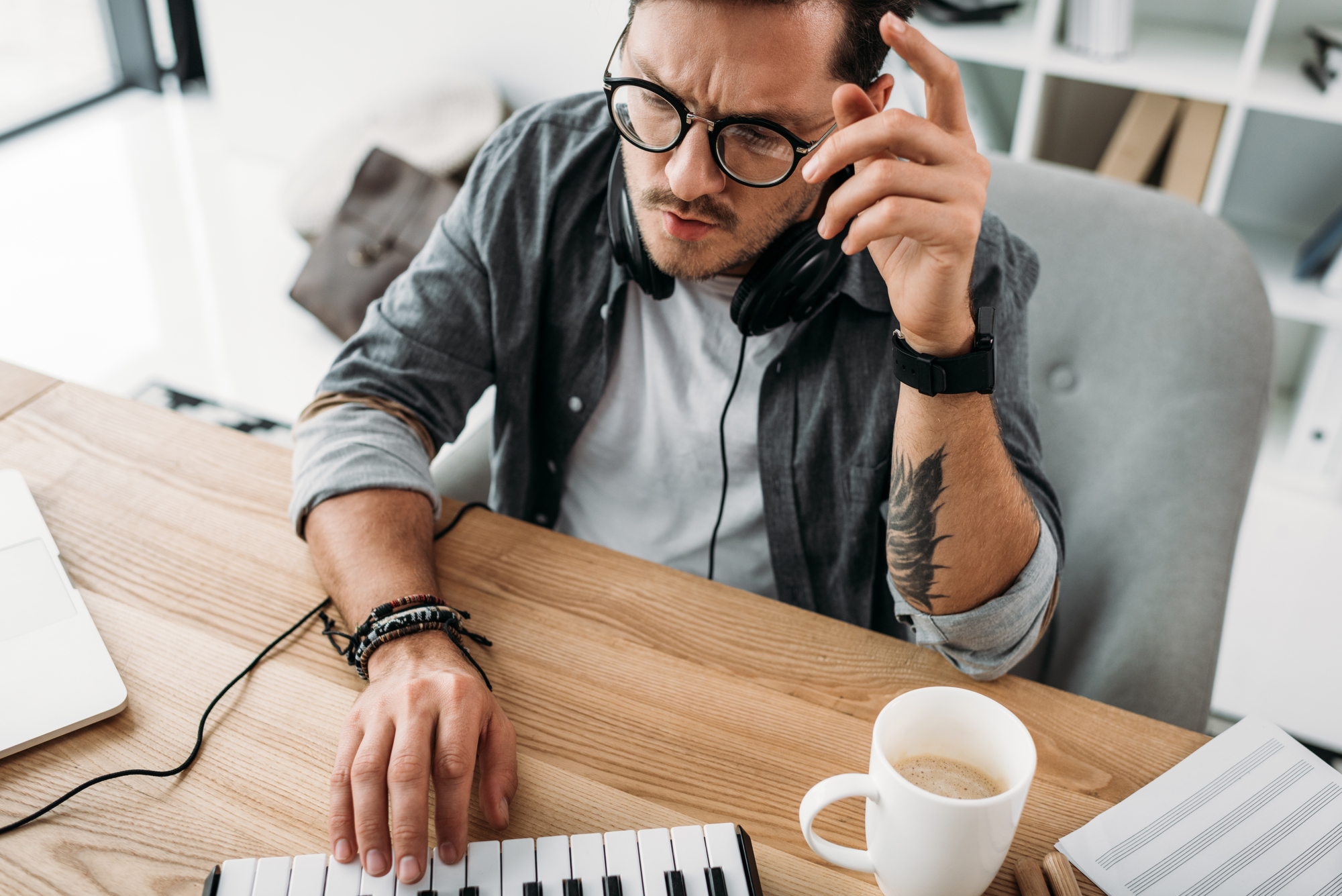 A man wearing glasses and headphones sits at a desk with a MIDI keyboard, working intently. His left hand is on the keyboard while his right hand is raised, appearing thoughtful. On the desk are a laptop, a cup of coffee, and a notepad.