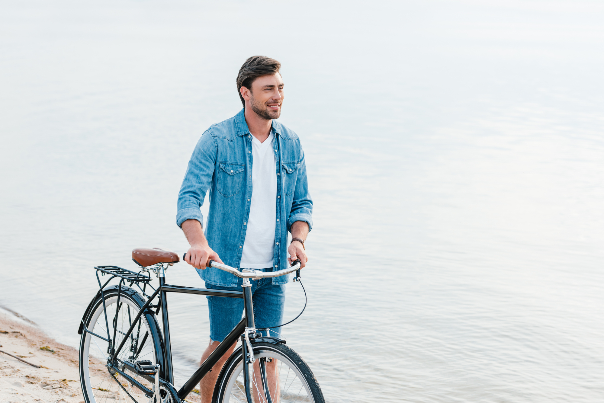 A man wearing a denim jacket and shorts stands on a sandy beach, holding a bicycle. The calm water of a lake or sea is visible in the background. He is looking into the distance and smiling.