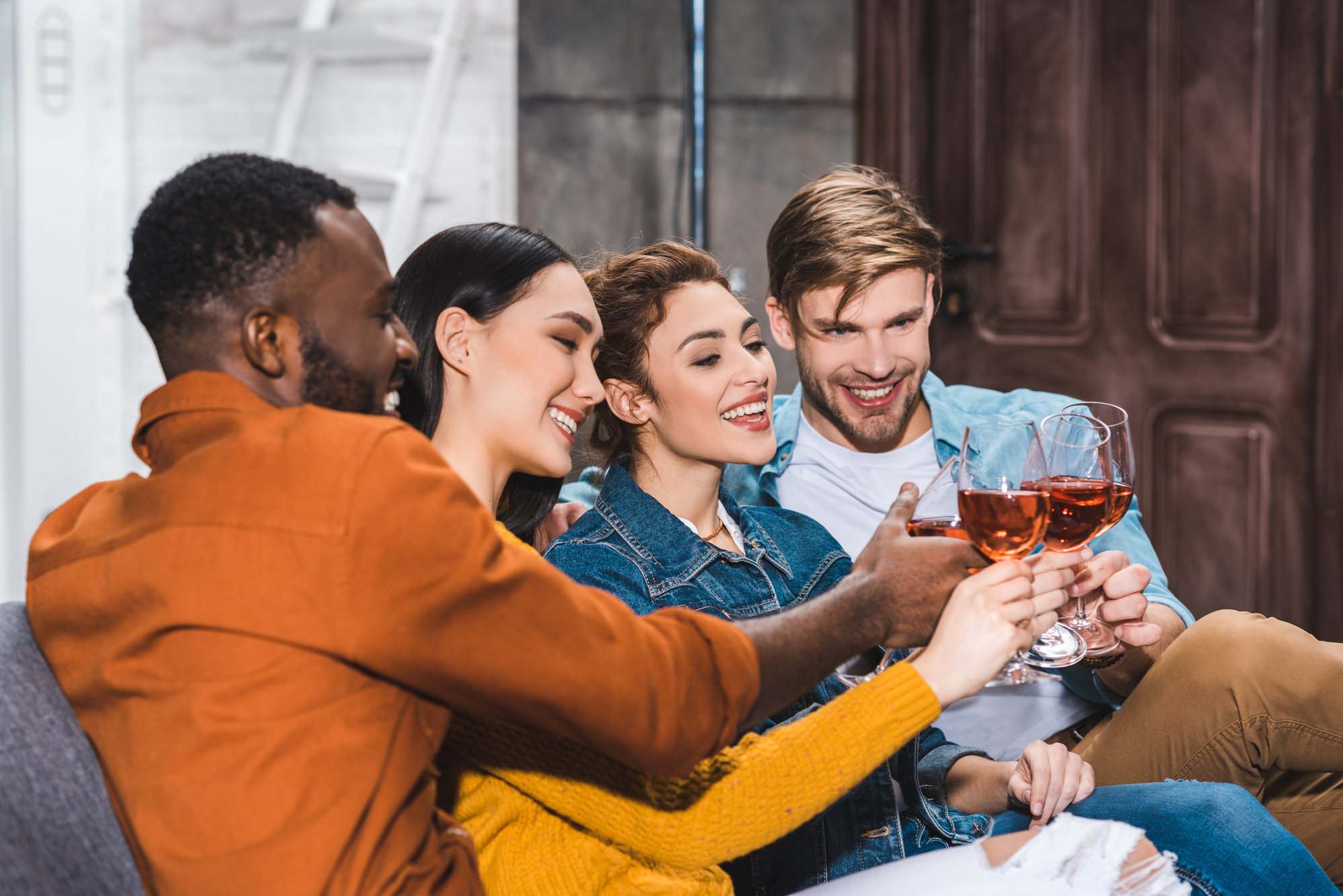 A group of four friends sitting closely together on a couch, smiling and raising their glasses in a toast. They are indoors, with a wooden door and white brick wall in the background. The mood appears joyful and celebratory, and the drinks seem to be wine.