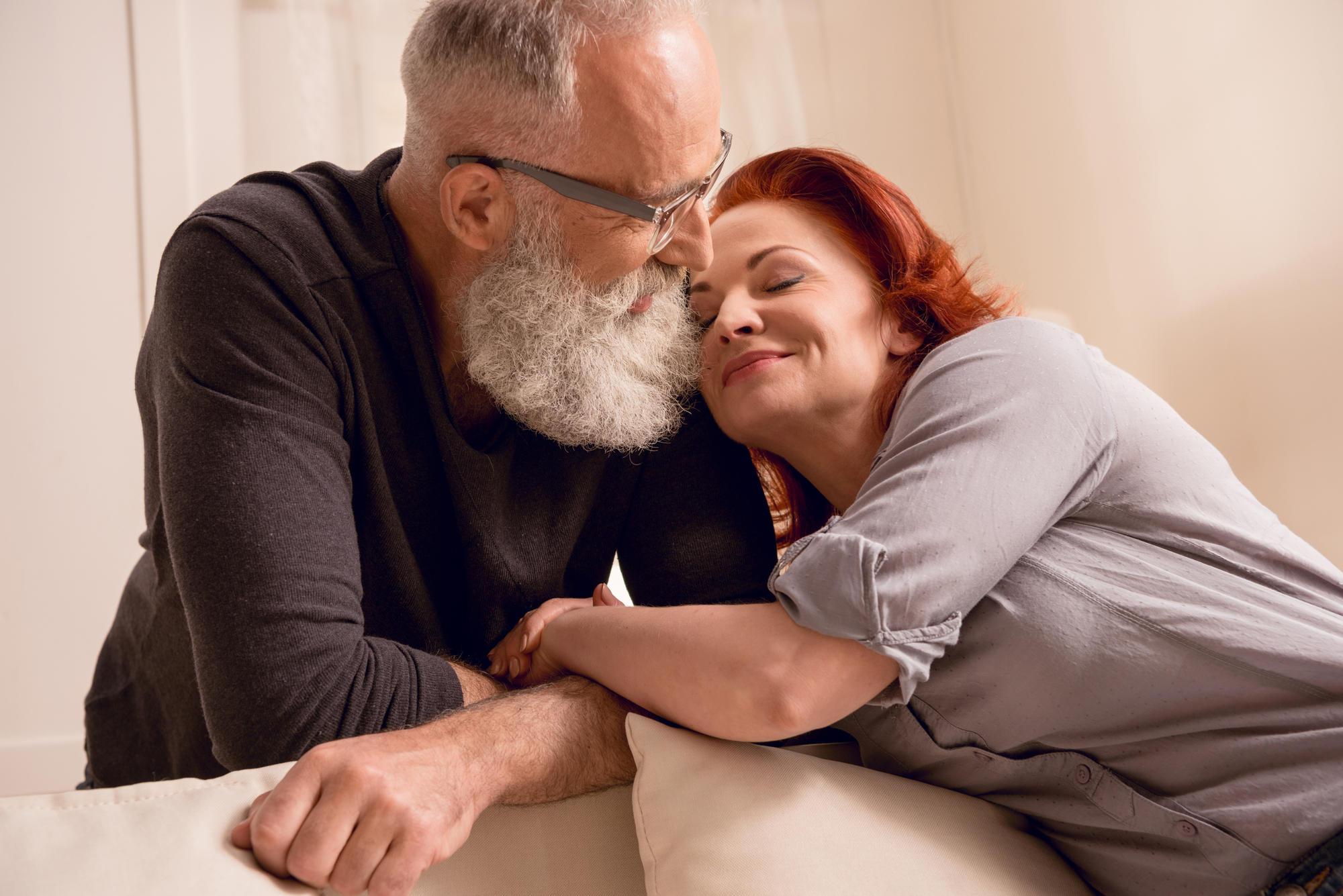 An elderly couple is sharing a tender moment indoors. The man, with a white beard and glasses, is leaning towards the woman who has red hair. She is smiling softly with her eyes closed, resting her head on his shoulder. Both are dressed in casual, comfortable clothing.