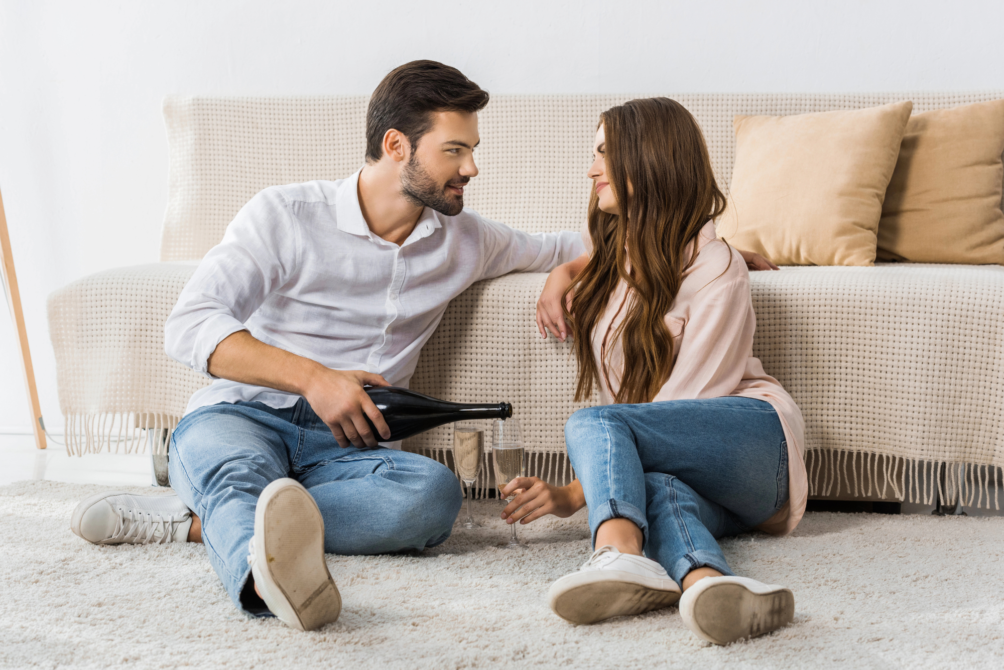 A man and woman sit on the floor in front of a beige couch. The man, holding a bottle, is pouring a drink into a glass held by the woman. Both are casually dressed in light-colored tops and jeans, and they are looking at each other, smiling.