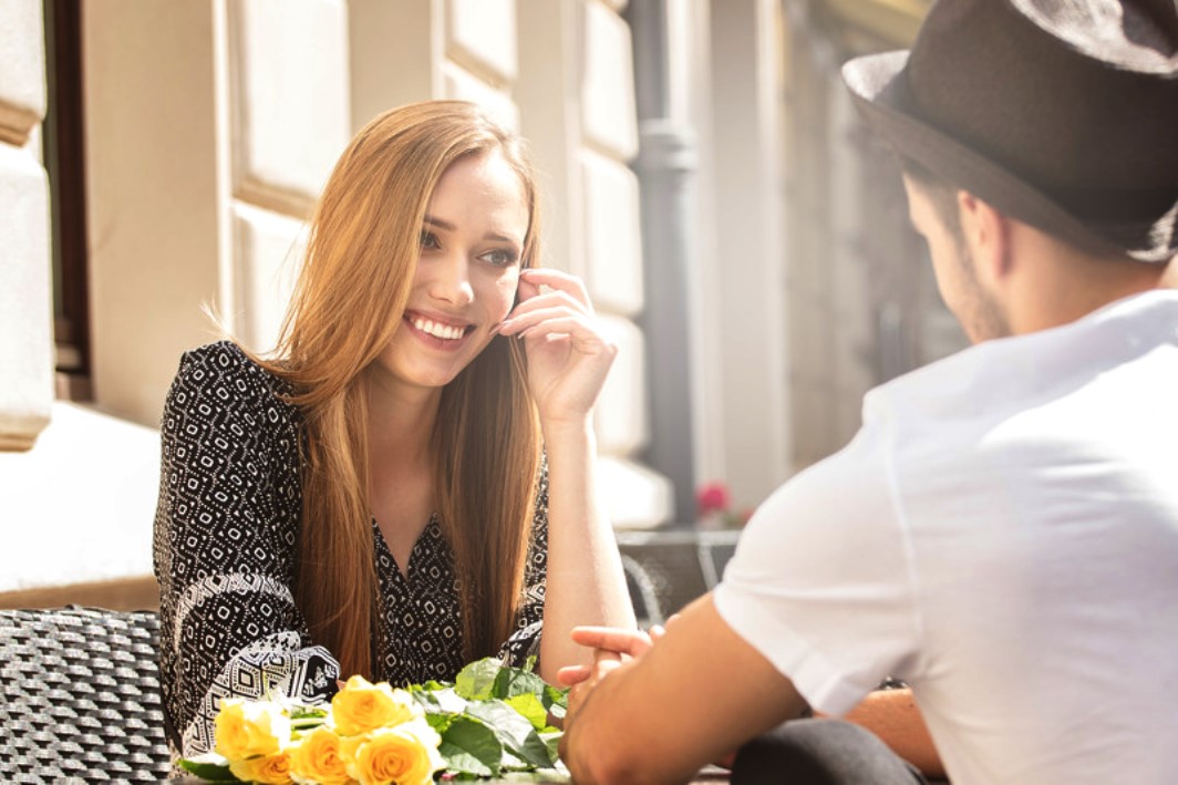 A young woman with long, light brown hair smiles while sitting at an outdoor café table with a bouquet of yellow roses in front of her. Across from her sits a man in a white t-shirt and black hat, partially turned away from the camera.