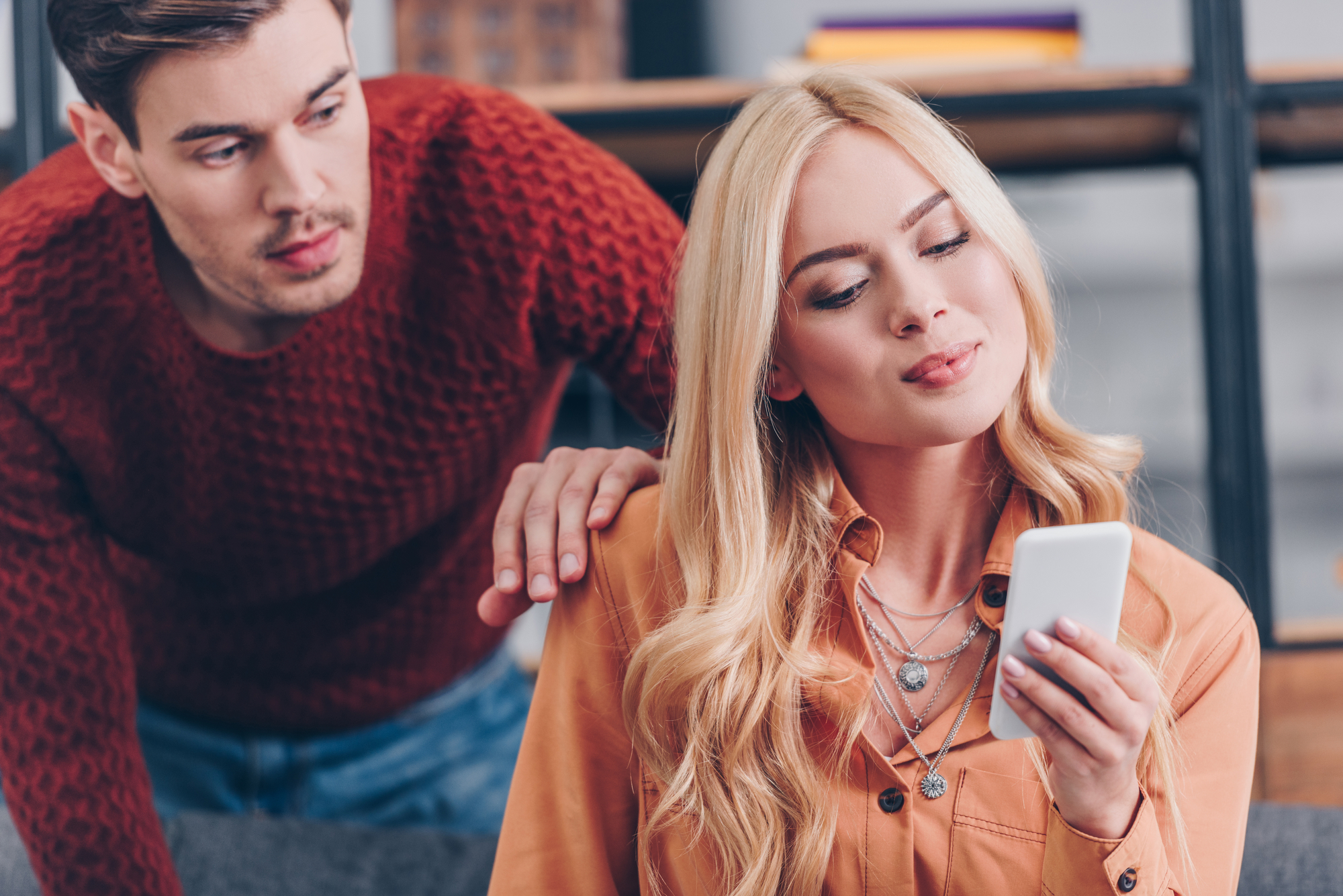 A blonde woman is sitting and looking at her phone with a small smile, while a man with short brown hair and beard, dressed in a red sweater, leans over her shoulder with a slightly curious expression. Shelves are visible in the background.