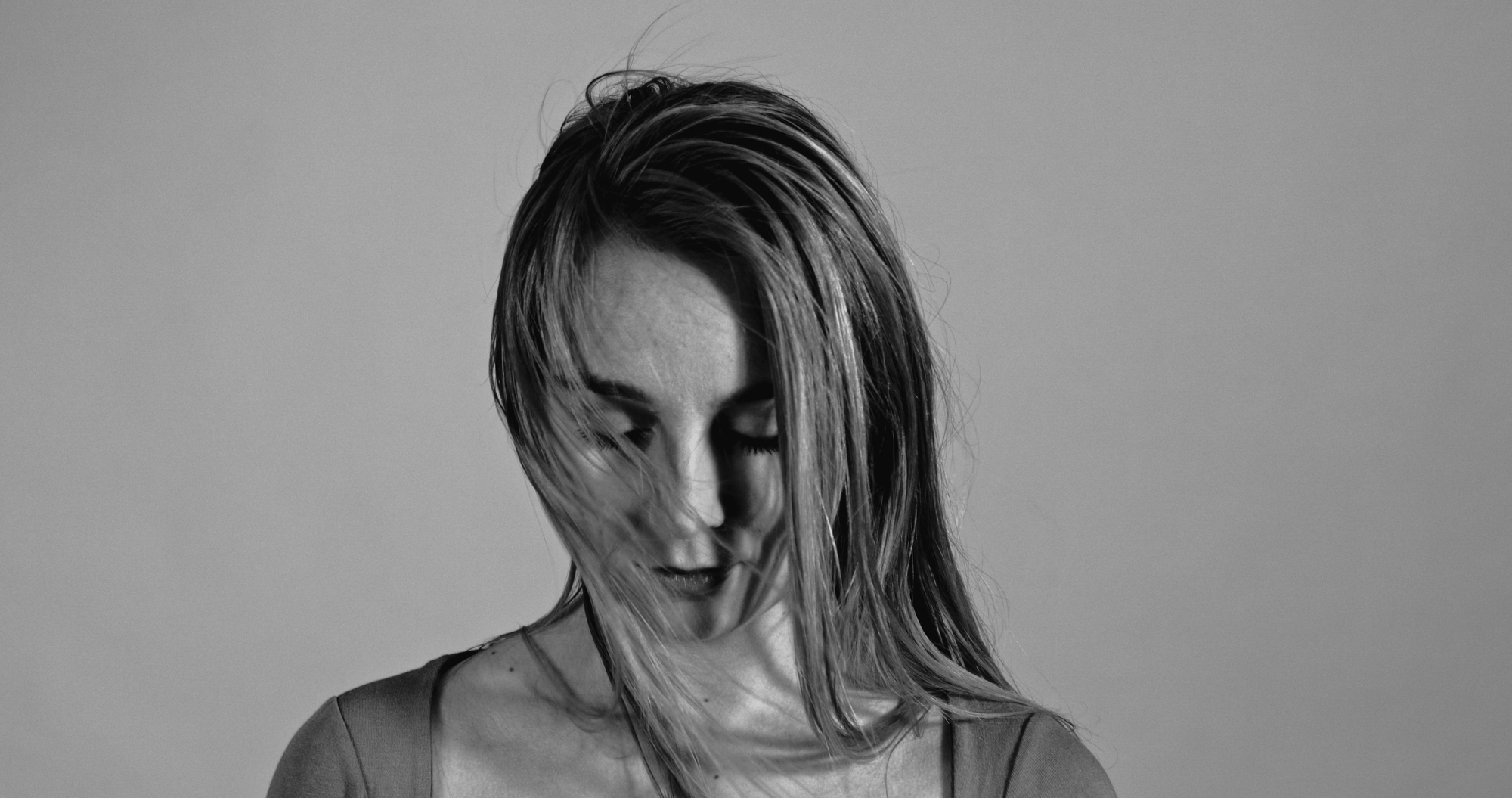 A grayscale image of a person with long hair, partially obscuring their face. Their head is tilted downward, with hair falling over their face, giving a contemplative or pensive expression. The background is plain.