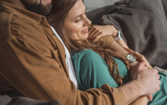 A couple relaxes on a couch. The person on the left in a brown jacket holds the person on the right, who has long hair and is wearing a teal shirt. Both appear content, and the person in teal is smiling with their head resting on the other's chest.