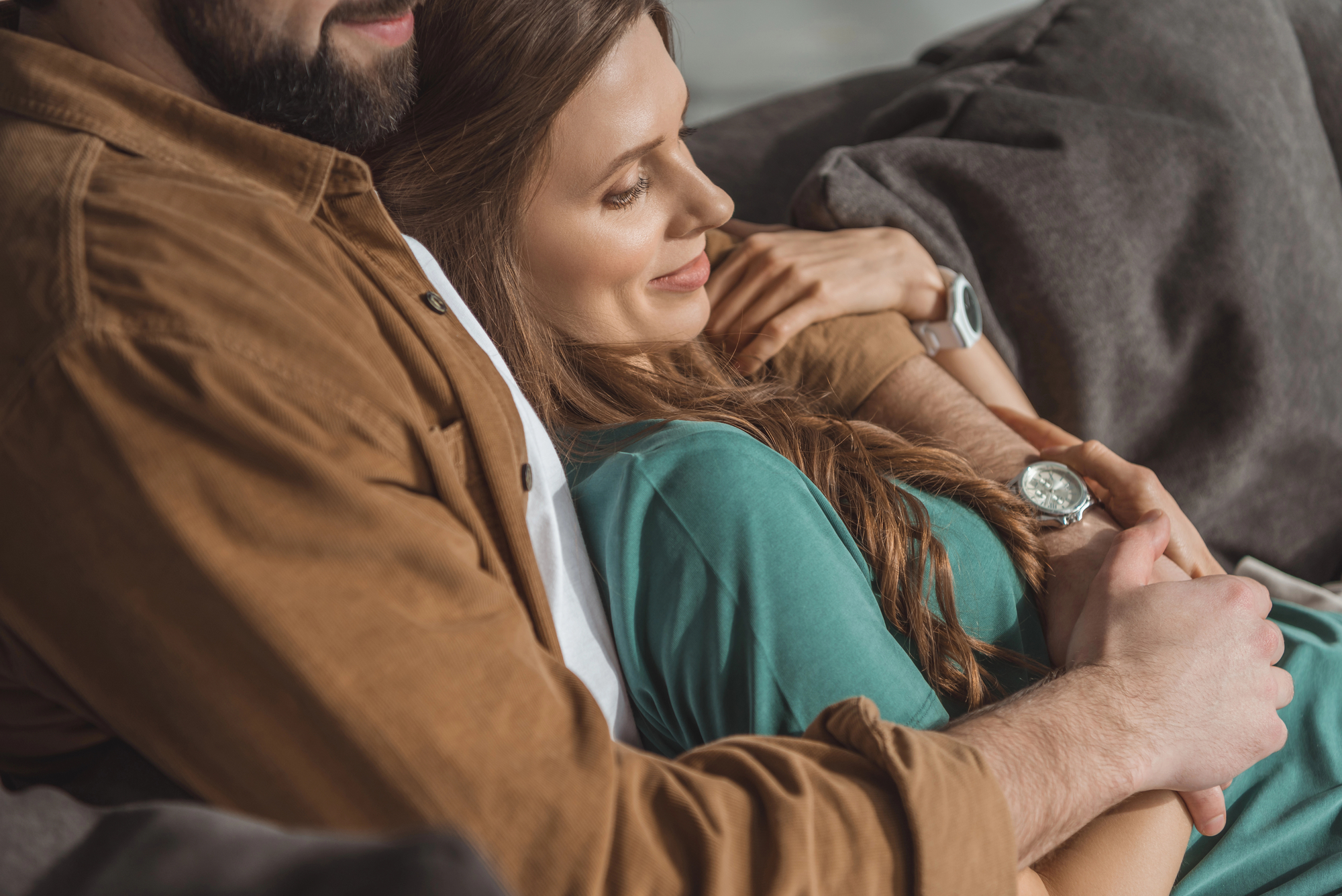 A couple relaxes on a couch. The person on the left in a brown jacket holds the person on the right, who has long hair and is wearing a teal shirt. Both appear content, and the person in teal is smiling with their head resting on the other's chest.