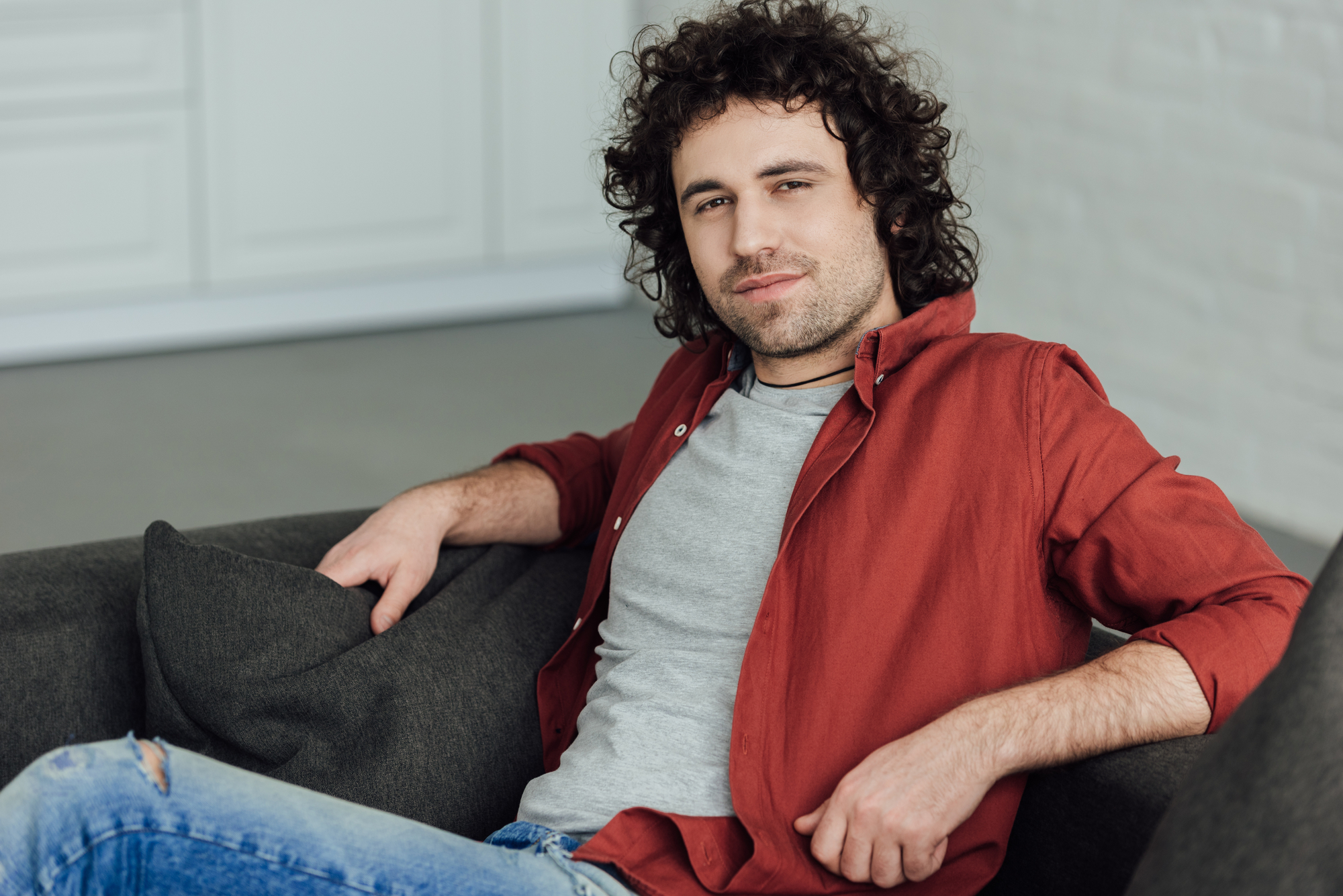 A young man with curly hair and a beard is sitting on a couch, leaning back with one arm resting on the backrest. He is wearing a red button-up shirt over a gray t-shirt and ripped blue jeans. He is looking at the camera with a relaxed expression.