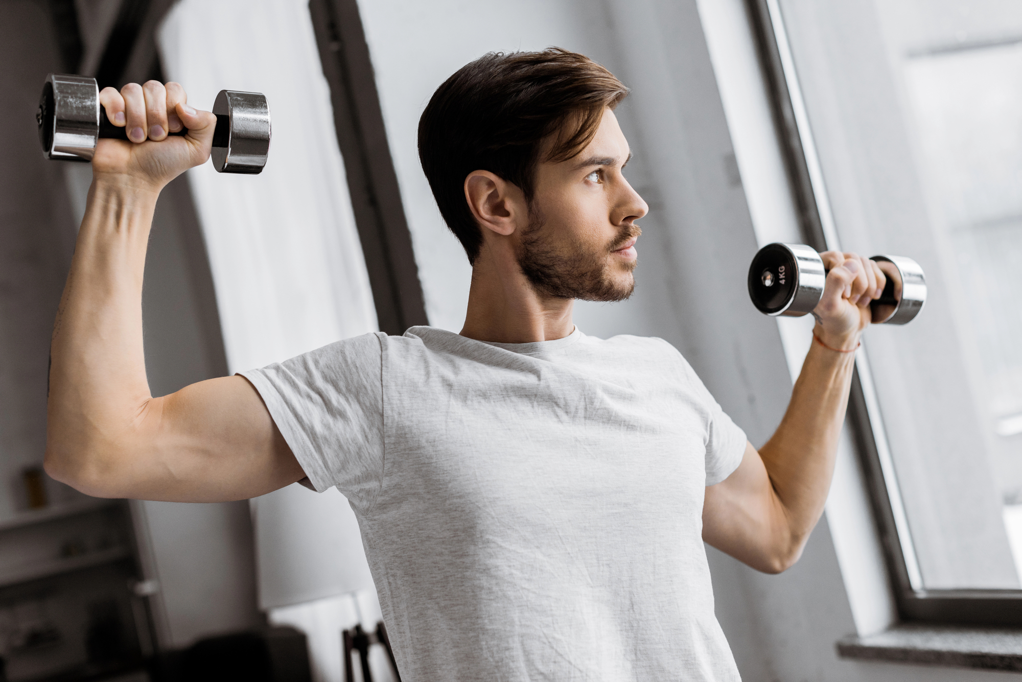 A man with short brown hair and a beard, wearing a white t-shirt, exercises by lifting two silver dumbbells, one in each hand, at shoulder height. He is standing near a large window in a well-lit room, looking focused towards his right side.