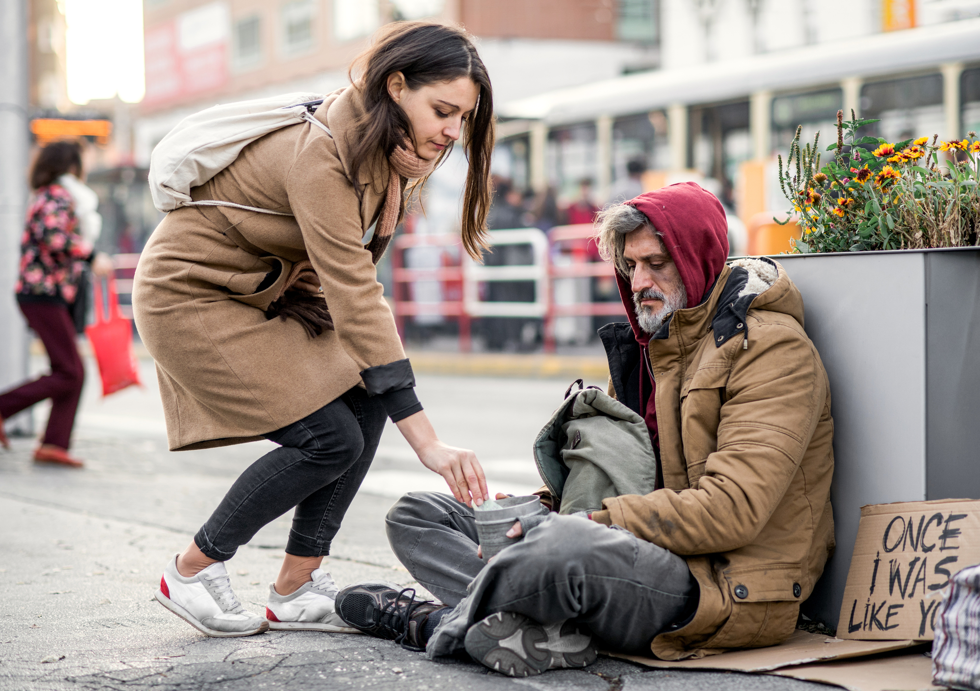 A woman in a tan coat kneels to give change to a homeless man sitting on the sidewalk. The man, wearing a red hoodie under a brown coat, has a sign beside him that reads "Once I was like you." A bus is visible in the background with people walking by.