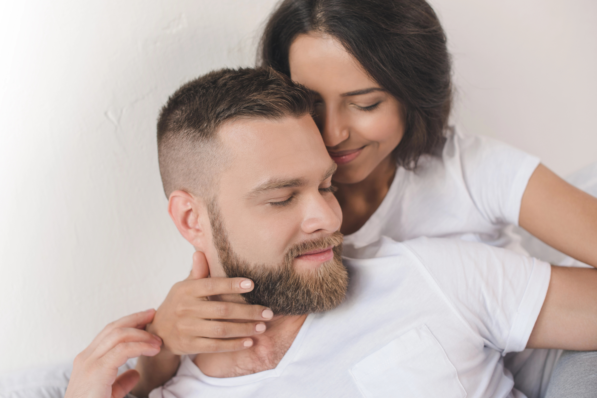 A couple wearing white shirts is lying down and cuddling. The man, with a beard and short hair, is leaning back with a content expression, while the woman, with shoulder-length hair, embraces him from behind, resting her head on his shoulder and smiling gently.