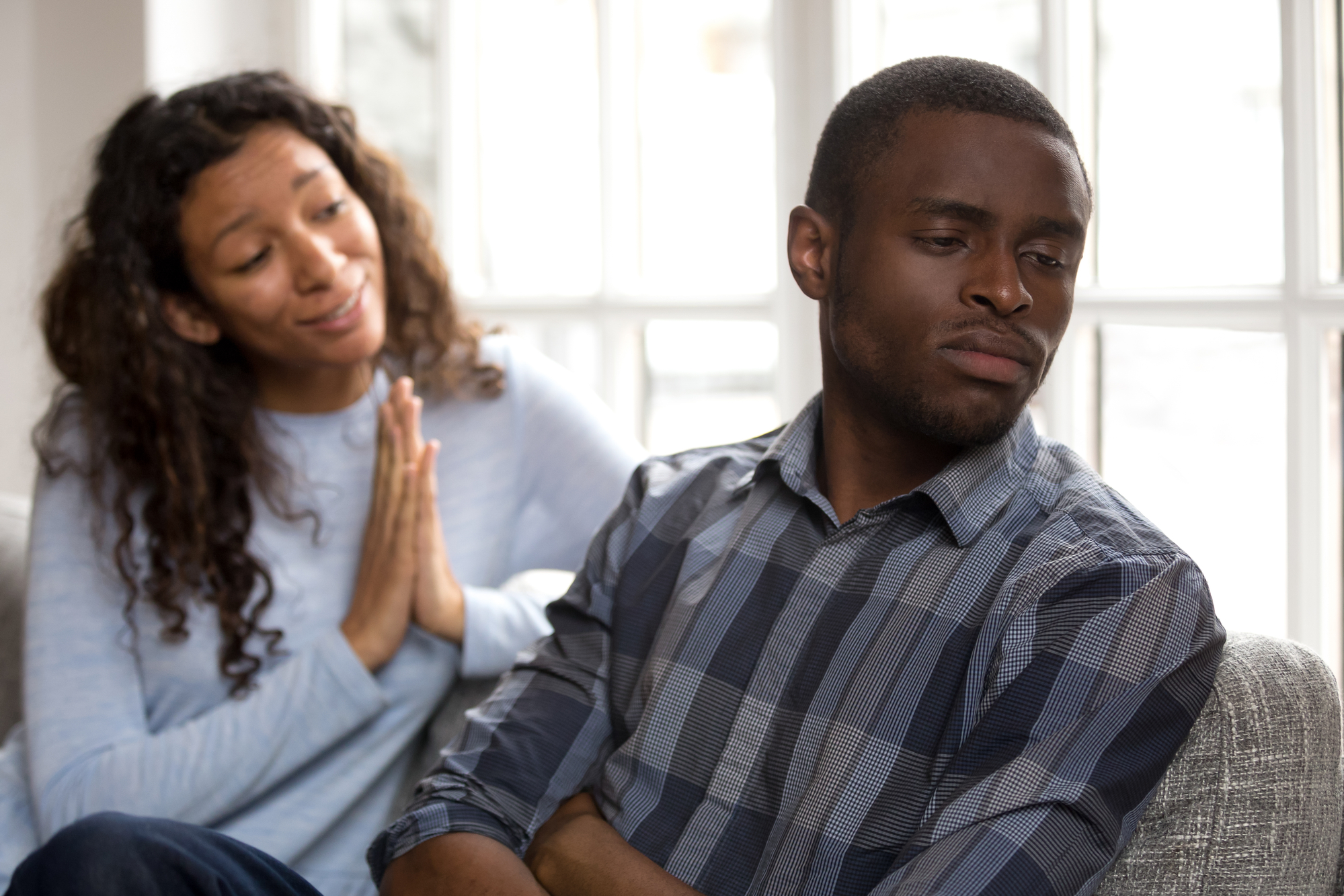 A woman with curly hair appears to be pleading or apologizing, her hands clasped together, while a man sitting next to her looks away with a stern or upset expression. They are indoors with a bright window in the background.