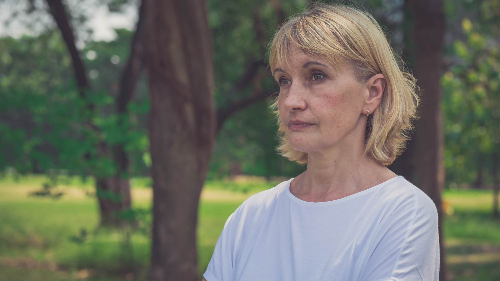 A middle-aged woman with short blonde hair and wearing a white shirt stands outdoors with her arms crossed. She has a serious expression and is looking off into the distance. The background features green trees and sunlight filtering through the leaves.