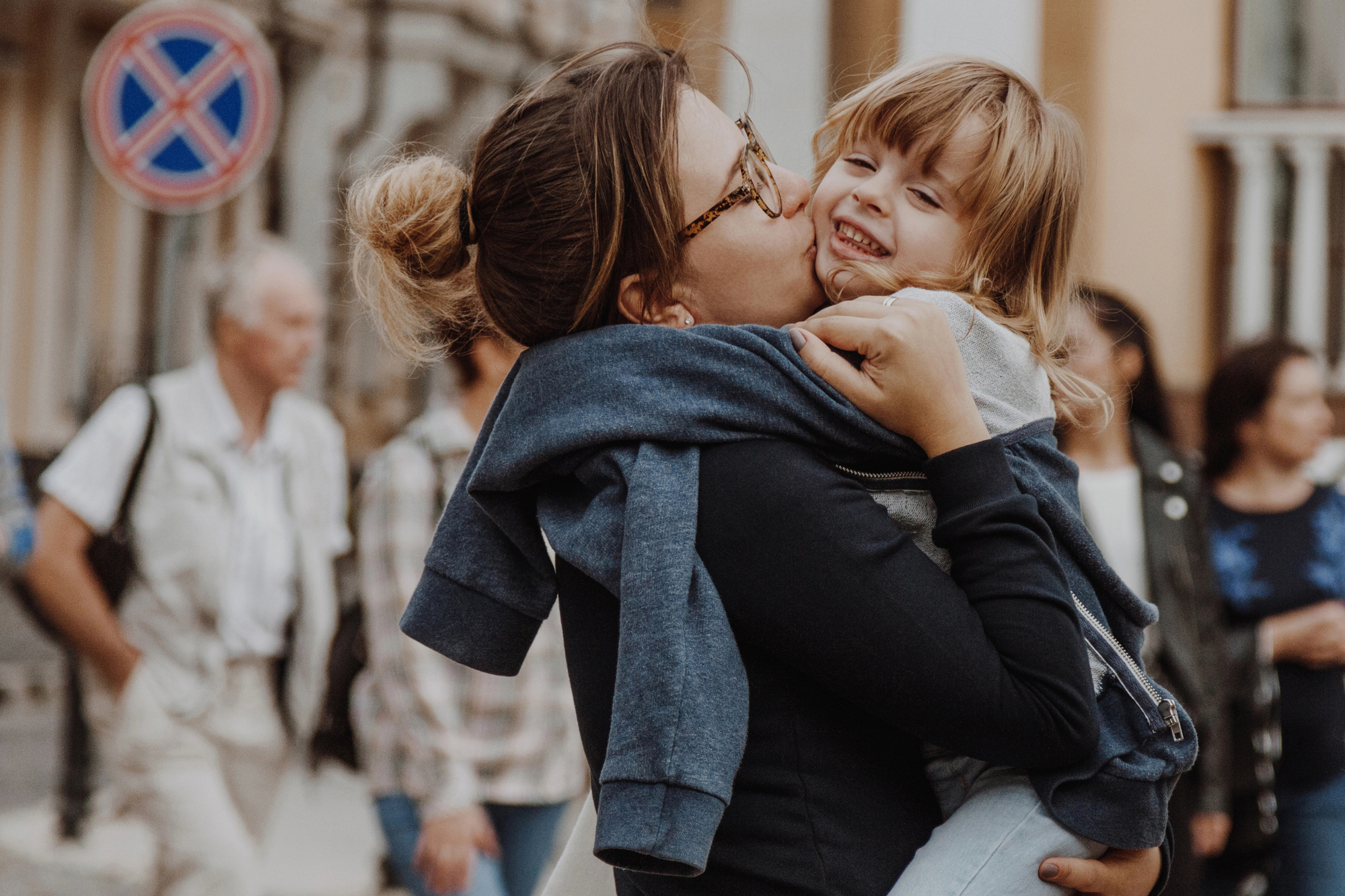 A woman with glasses kisses a smiling child on the cheek while holding them in her arms on a busy street. The child is giggling and wearing a blue hoodie. In the background, there are several people walking and a no parking sign is visible.