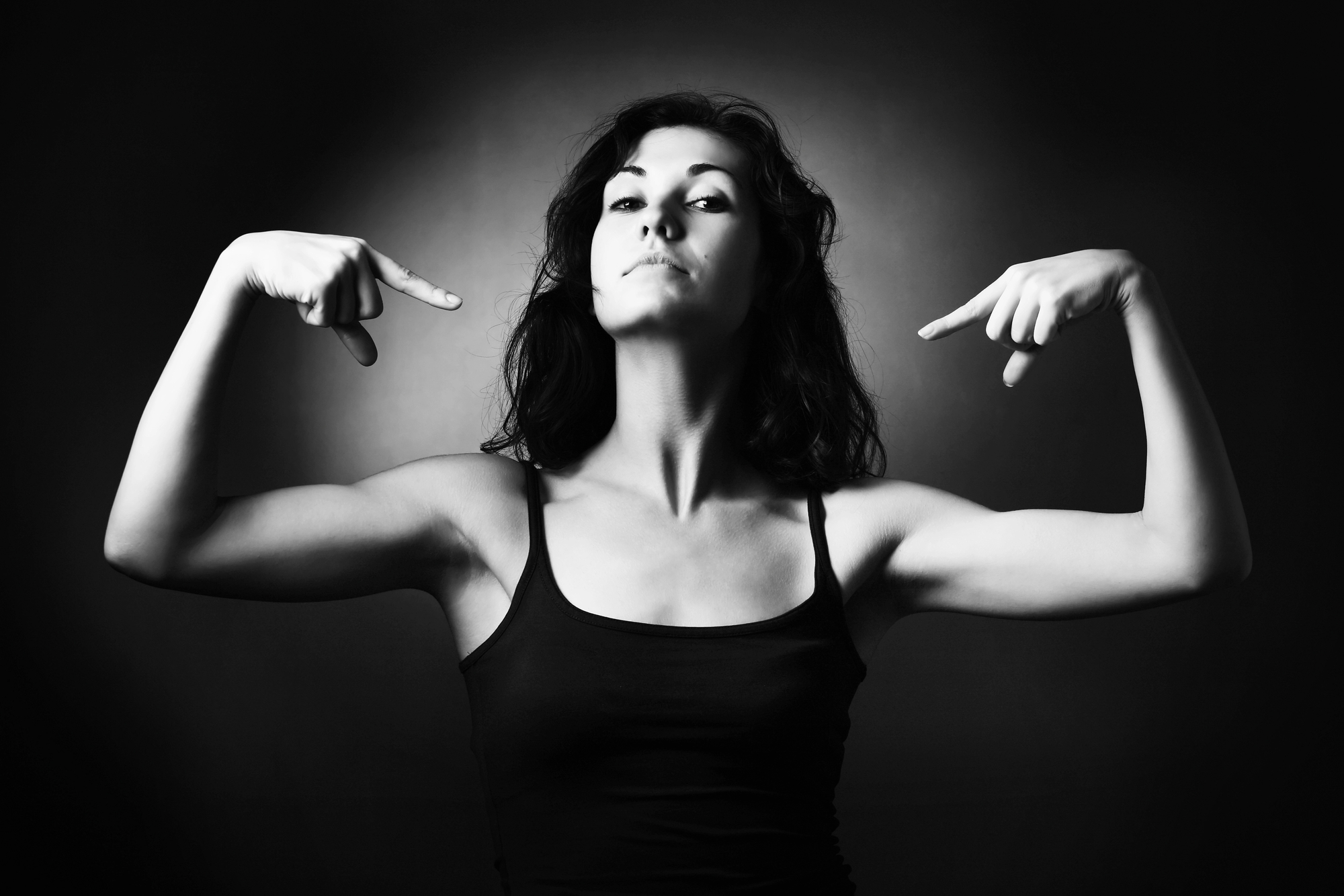 Black and white image of a woman with medium-length dark hair, wearing a tank top, striking a confident pose. She has her head raised, with both arms bent at the elbows and pointing at herself with her index fingers. The background is dark and gradient.