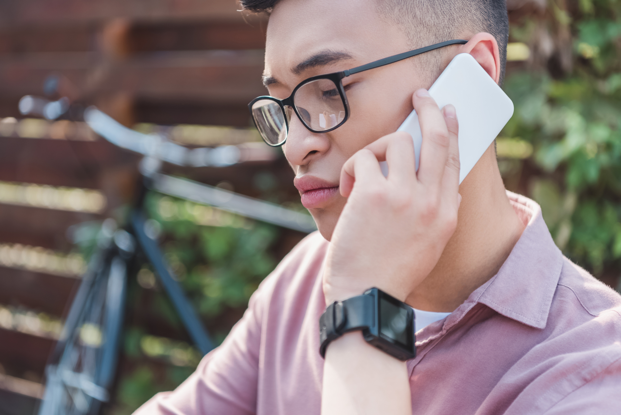 A person with short hair and glasses is holding a smartphone to their ear, appearing to be in a conversation. They are wearing a pink shirt and a smartwatch. In the background, a parked bicycle is visible, along with some greenery and a wooden fence.