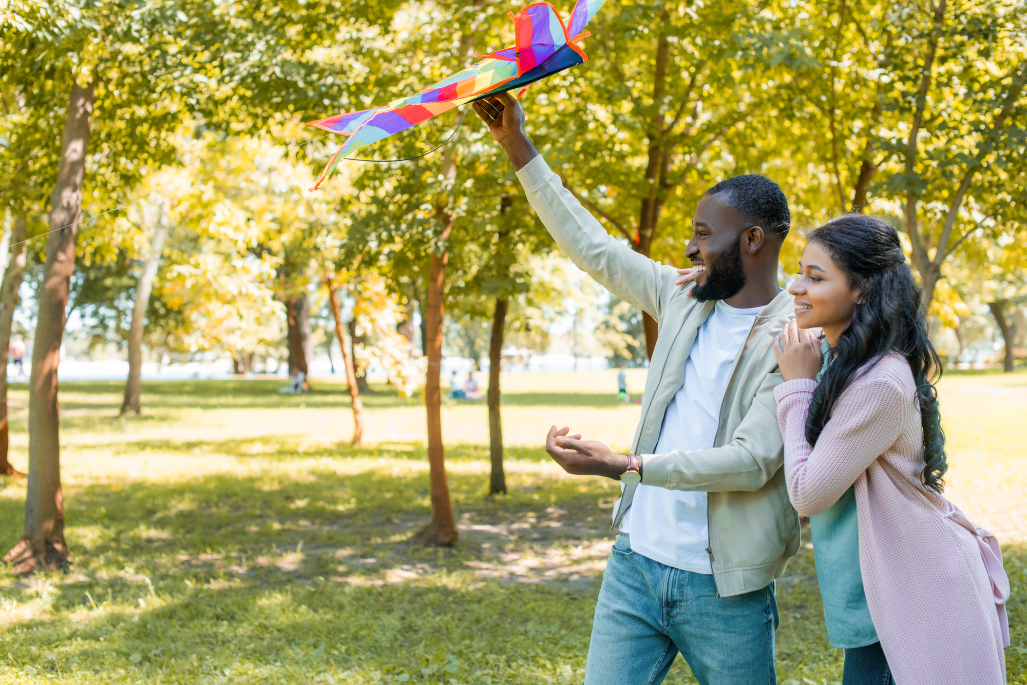 A man and woman are smiling as they get ready to fly a colorful kite in a sunlit park. The man holds the kite above his head while the woman stands close, looking at the kite. Trees with green foliage surround them, and soft sunlight filters through the branches.