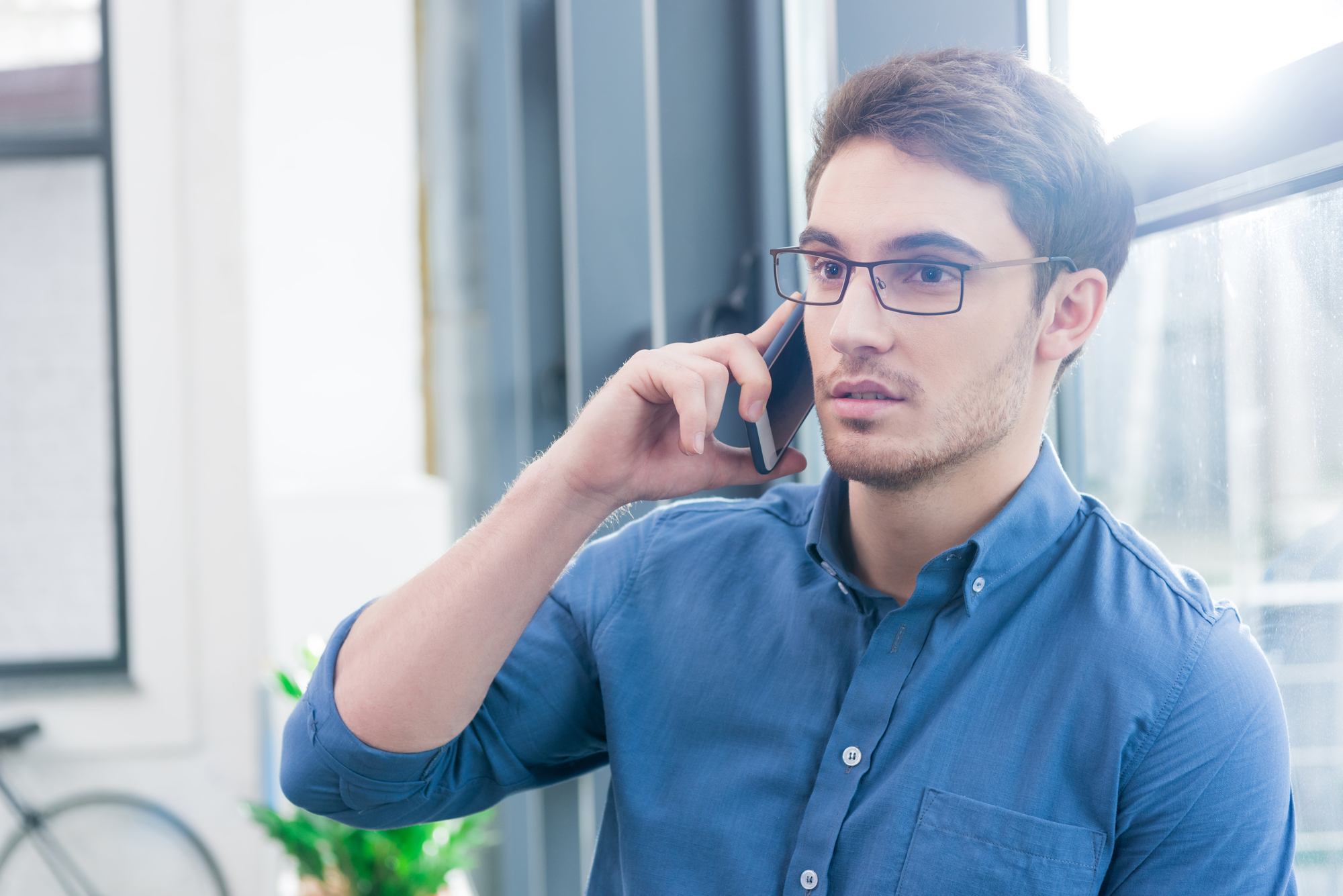 A man with short brown hair and glasses is talking on a smartphone. He is wearing a blue button-down shirt and is standing near a window with natural light coming in. There is a blurry view of a bicycle and a plant in the background.
