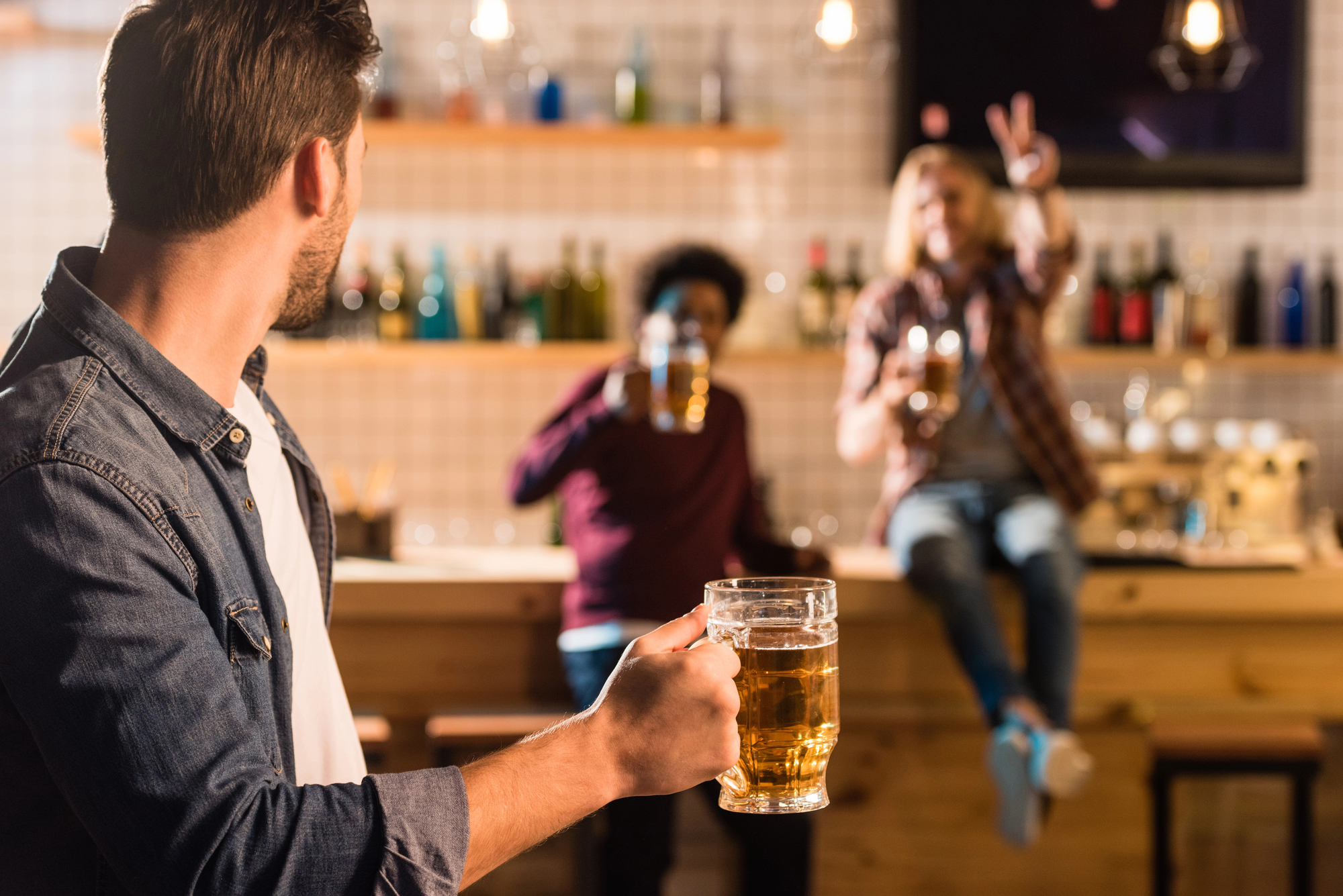 A man holding a mug of beer looks towards two people in the background who are also holding drinks. One of them is sitting on a bar counter making a peace sign with their fingers. The scene is in a warm, softly lit bar with shelves of bottles behind them.