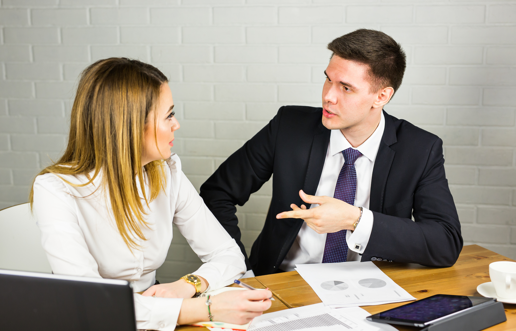 A man and a woman, both in business attire, are seated at a wooden table in a professional setting. They are engaged in a discussion, with documents and a laptop in front of them. The man is gesturing with his hand, while the woman listens attentively.