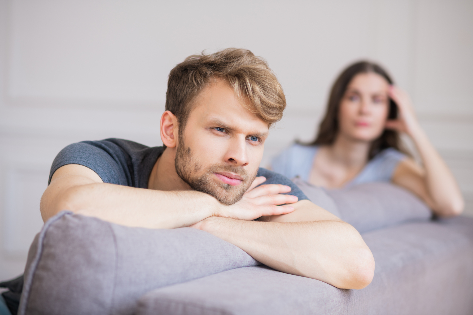 A man with a thoughtful expression leans on a gray couch with his arms crossed. In the background, a woman sits with her hand resting on her head, appearing concerned or pensive. Both are indoors in a softly lit room with light-colored walls.