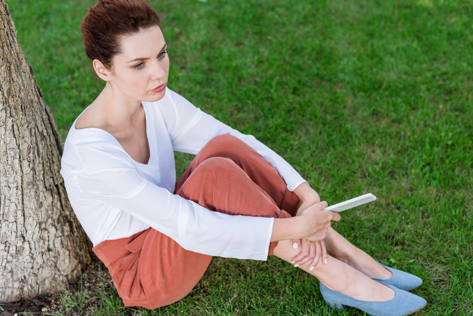 A woman with reddish-brown hair sits on the grass, leaning against a tree. She is wearing a white long-sleeve top, orange pants, and blue shoes, and holds a smartphone in her hand. She looks pensive and deep in thought.