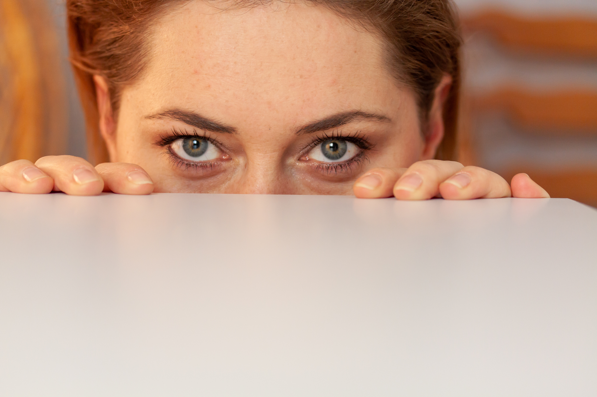 Close-up of a person with light-colored eyes peeking over the edge of a white surface. Only the top half of their face and hands gripping the surface are visible. The background is blurred, suggesting an indoor setting.