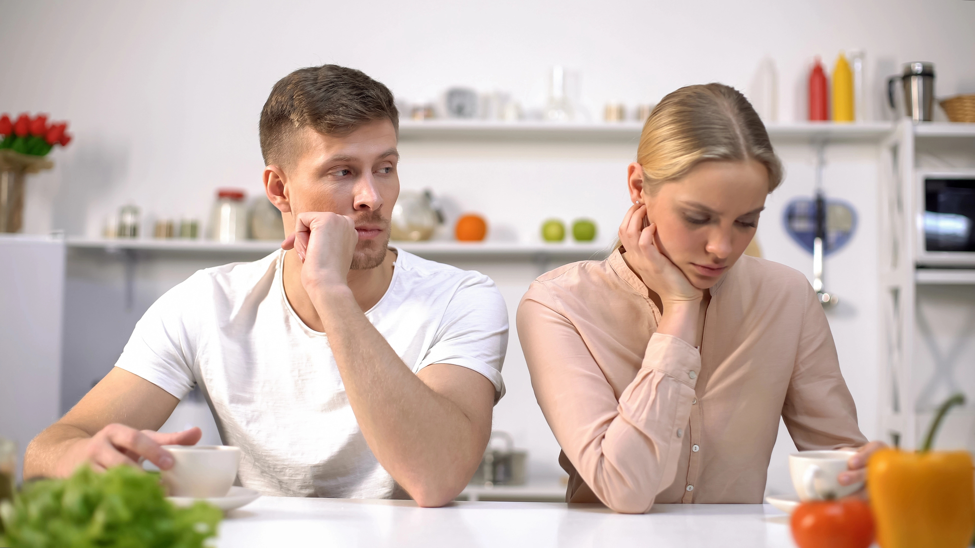 A man and woman sit at a table in a kitchen, both resting their chins on their hands with thoughtful, concerned expressions. The man looks at the woman, while she gazes downward. Various kitchen items are visible on shelves in the background.