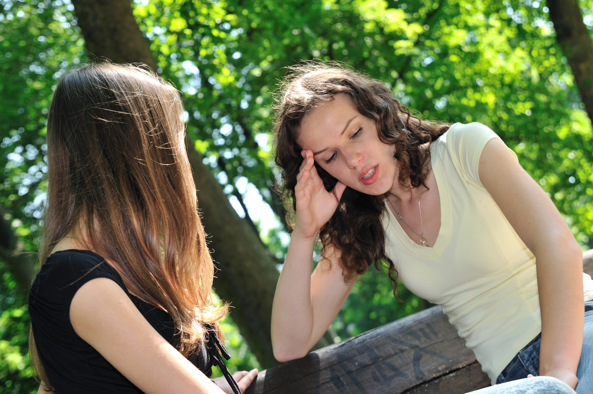 Two women are sitting on a bench in a park with green trees in the background. One woman with curly hair holds her head as if in distress or discomfort, while the other woman, with long straight hair, sits beside her, appearing to be listening or talking to her.