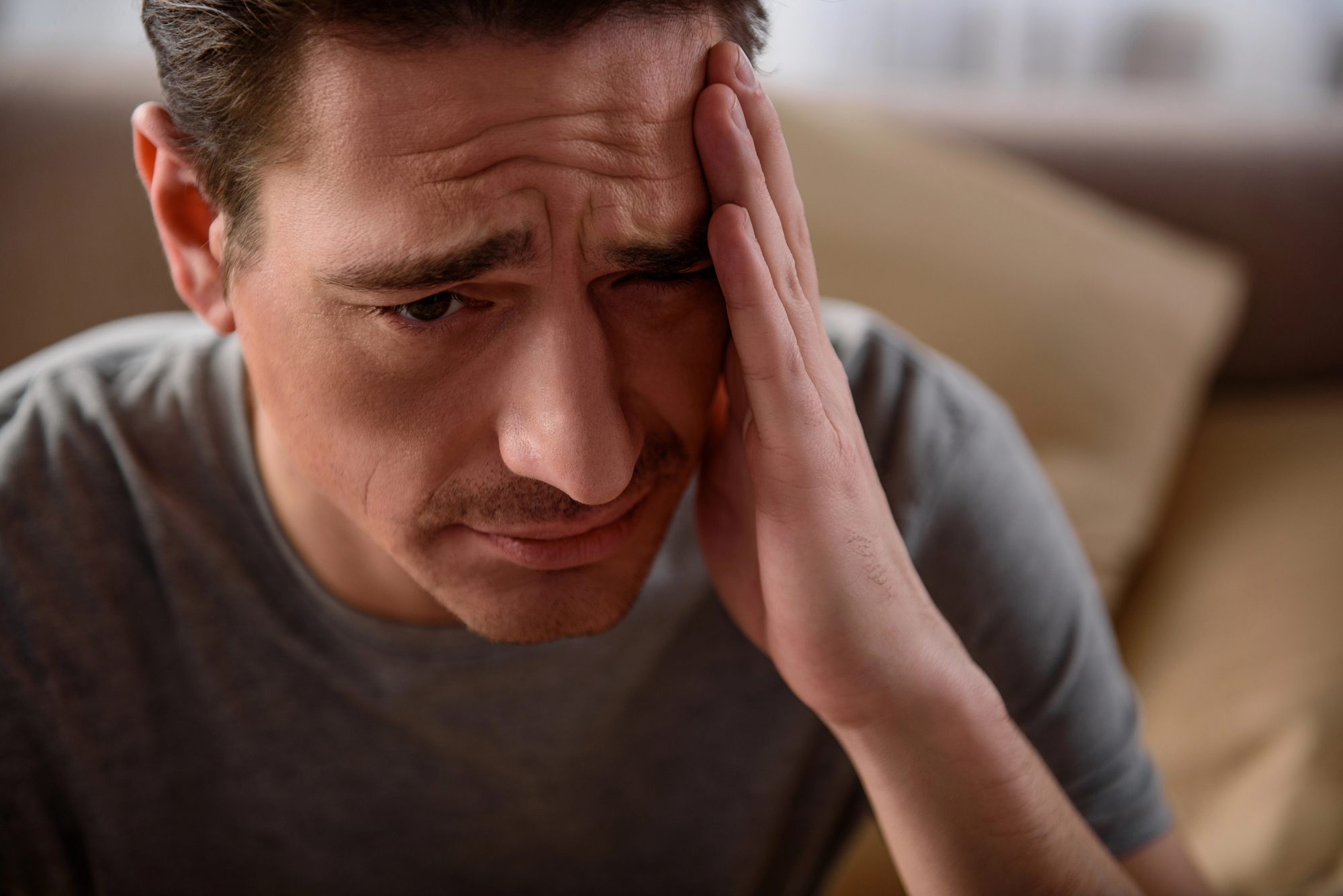 A man is sitting on a couch, looking distressed. He is wearing a grey t-shirt and is holding his hand to his forehead, appearing to have a headache or be in deep thought. The background is blurred, with neutral-colored cushions visible behind him.