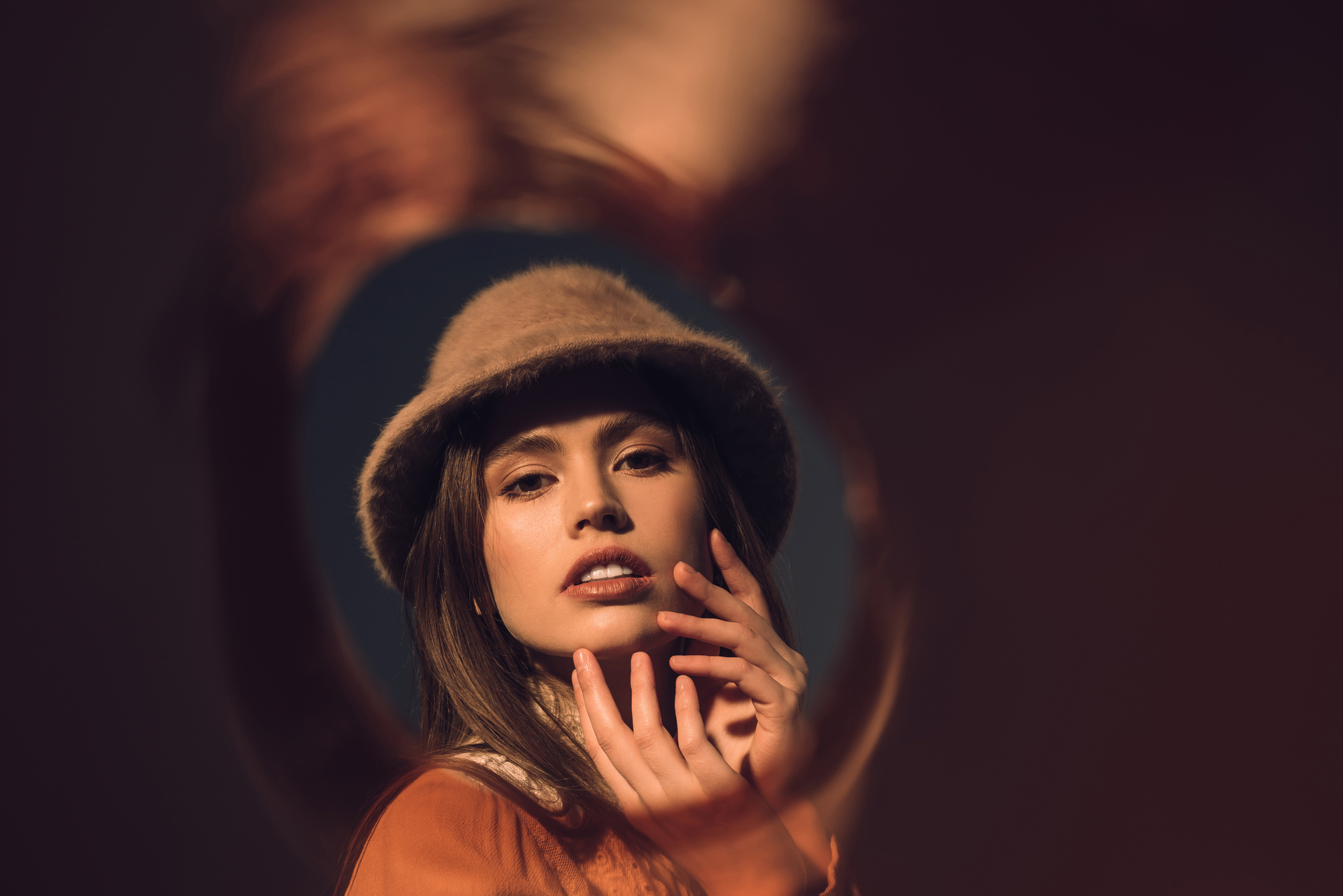 A woman wearing a brown hat and an orange shirt poses with her hands touching her face. The image has a warm, blurred vignette effect around the edges, drawing focus to her serene expression and subtle smile. The dark background contrasts with the warm tones.