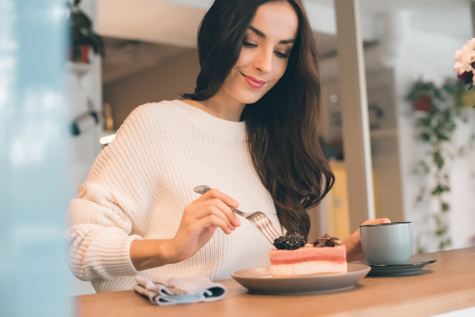 A woman with long dark hair sits at a wooden table, smiling while using a fork to eat a slice of cake topped with berries. She wears a white sweater and holds a cup with her other hand. A blue napkin rests on the table beside her. The background is softly blurred.