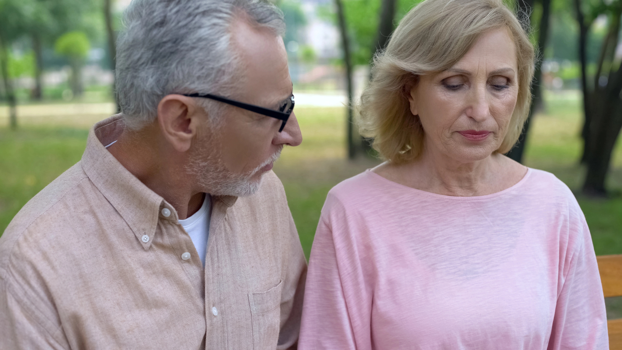 An older man with gray hair and glasses, wearing a light brown shirt, looks at an older woman with shoulder-length blonde hair, wearing a light pink shirt. They appear to be sitting on a bench in a park, with trees and greenery in the background.