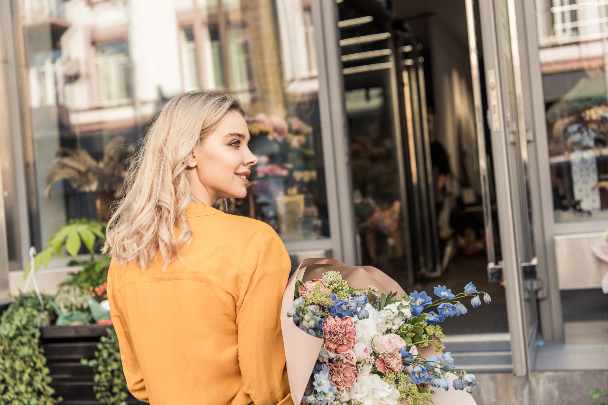 A woman with blonde hair, wearing a mustard yellow jacket, is smiling while holding a bouquet of colorful flowers. She is standing in front of an open storefront with plants and flowers visible within and outside the shop.